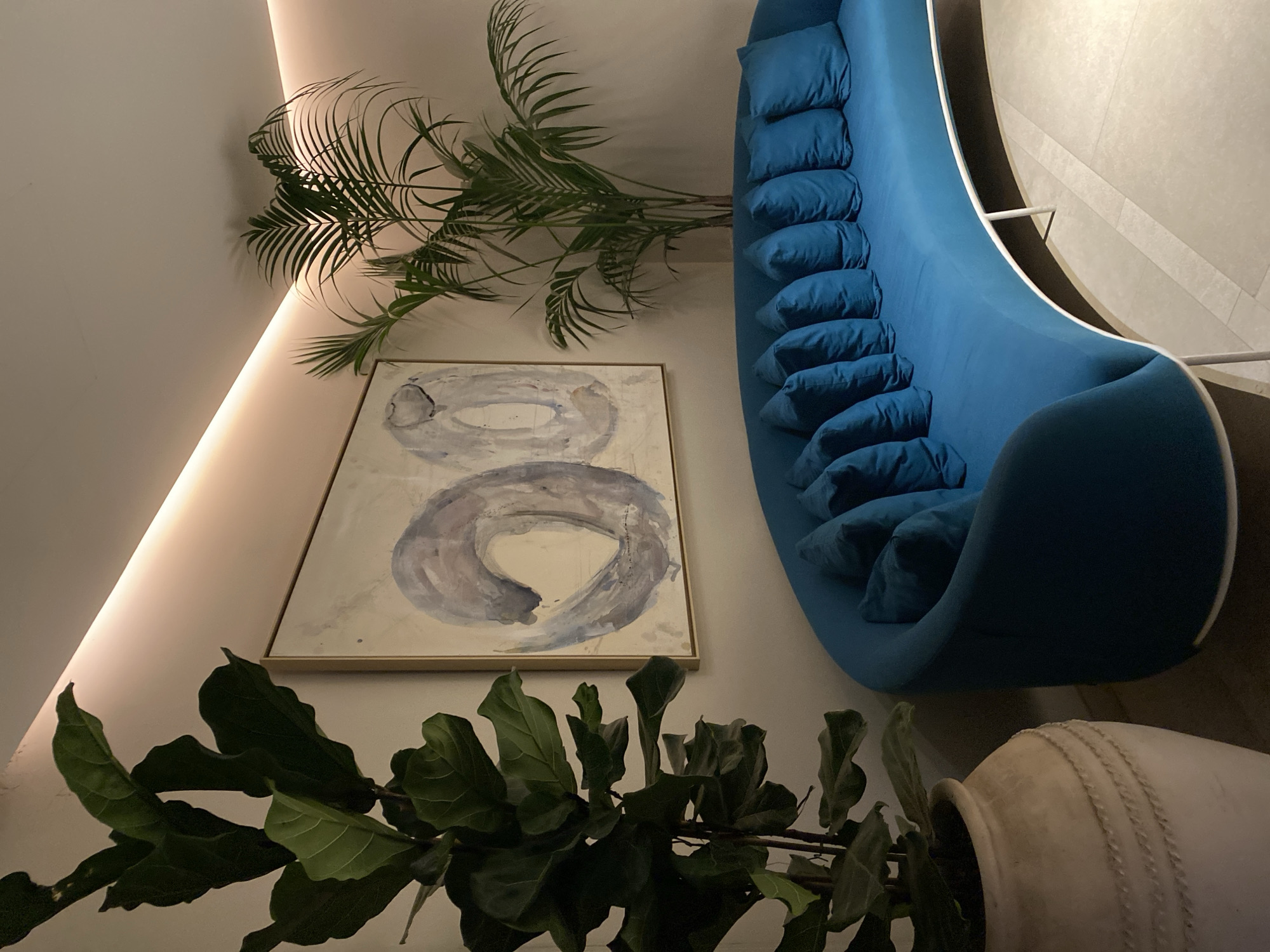 This image depicts a contemporary interior scene, likely in a hotel lobby or a modern living space, in Estepona, Spain. The focal point is a vibrant, curved, teal-blue sofa with multiple matching cushions. To the left of the sofa, a large potted fiddle-leaf fig plant stands tall in a textured, light-colored pot. Behind the sofa, a piece of abstract art with blue and gray brushstrokes is displayed on a pale wall. To the right of the artwork, a palm plant adds a touch of greenery to the space. The room is illuminated by subtle, recessed lighting along the ceiling, casting a warm glow and creating soft shadows. The flooring appears to be large, light-colored tiles. There are no people visible in the image, and the scene is static and composed.