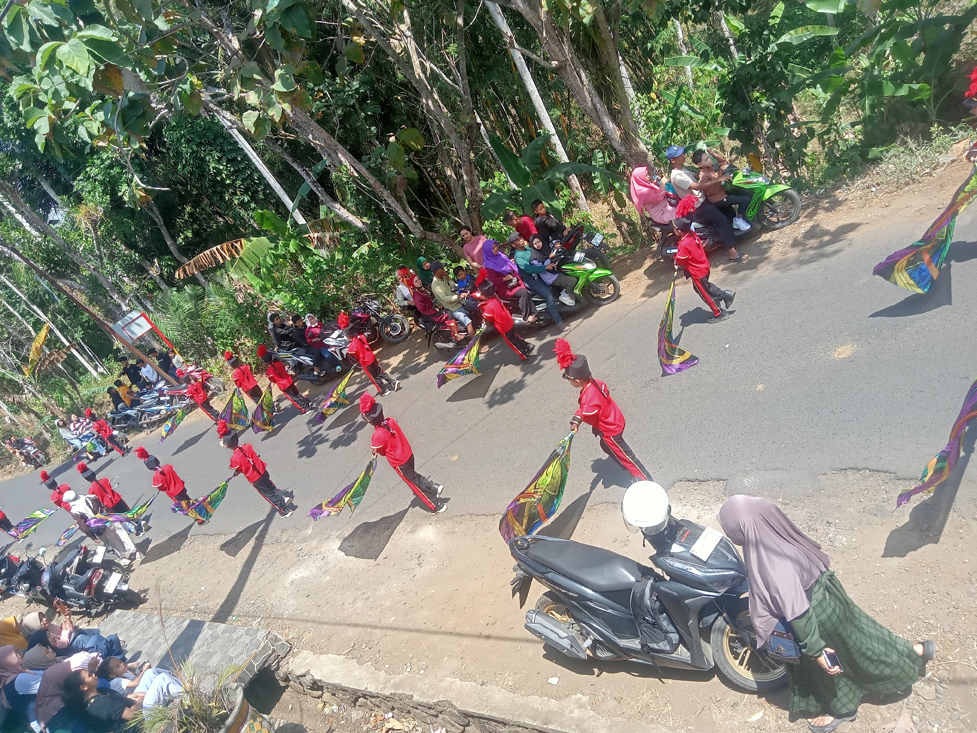In the city of Pule, Indonesia, a group of individuals in matching red uniforms with black trim and hats are participating in a parade or performance on a paved road. They are carrying colorful, patterned flags that are spread out as they move. The road is lined with trees and vegetation on the right side. On the left side of the road, a group of spectators are gathered, many sitting or standing near parked motorcycles. Some people are seated on motorcycles, while others are standing or sitting on the ground. A few motorcycles are parked along the side of the road. In the foreground, a black motorcycle with a white helmet on its seat is parked. The sky is visible, suggesting it is daytime with sunlight casting shadows.
