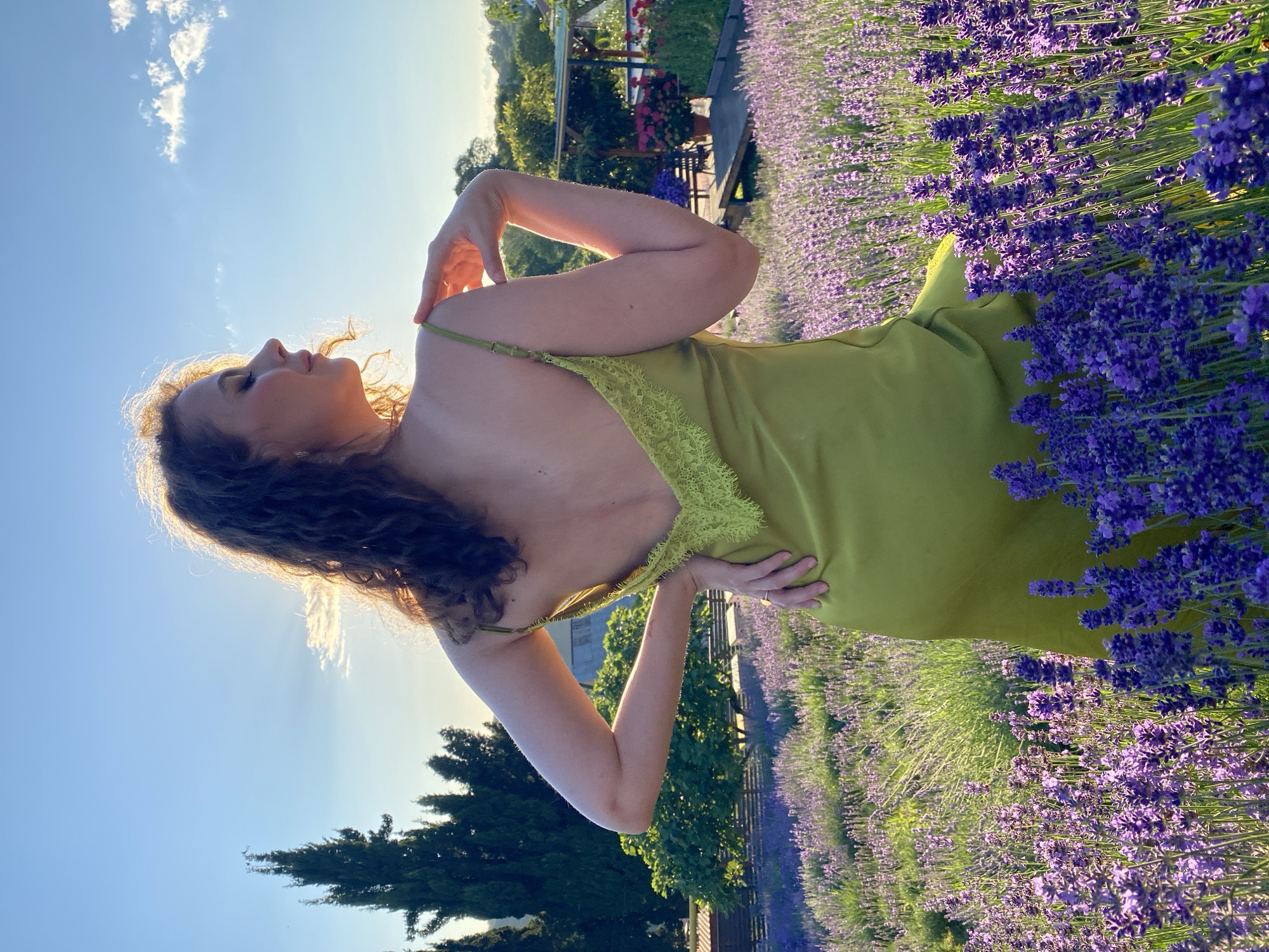 A woman with curly brown hair is posing in a field of lavender. She is wearing a lime green slip dress with lace detailing on the straps. Her arm is raised, with her hand touching her neck, and her other hand is resting on her hip. The setting appears to be a lavender farm, with rows of purple flowers stretching into the distance. Trees and a wooden structure are visible in the background. The sun is setting, casting a warm glow on the scene and creating long shadows. The sky is a clear blue with a few wispy clouds. The woman's pose and the lighting suggest a moment of relaxation and enjoyment. The location is Verkhneye Uchdere, Russia. There is no visible text in the image.
