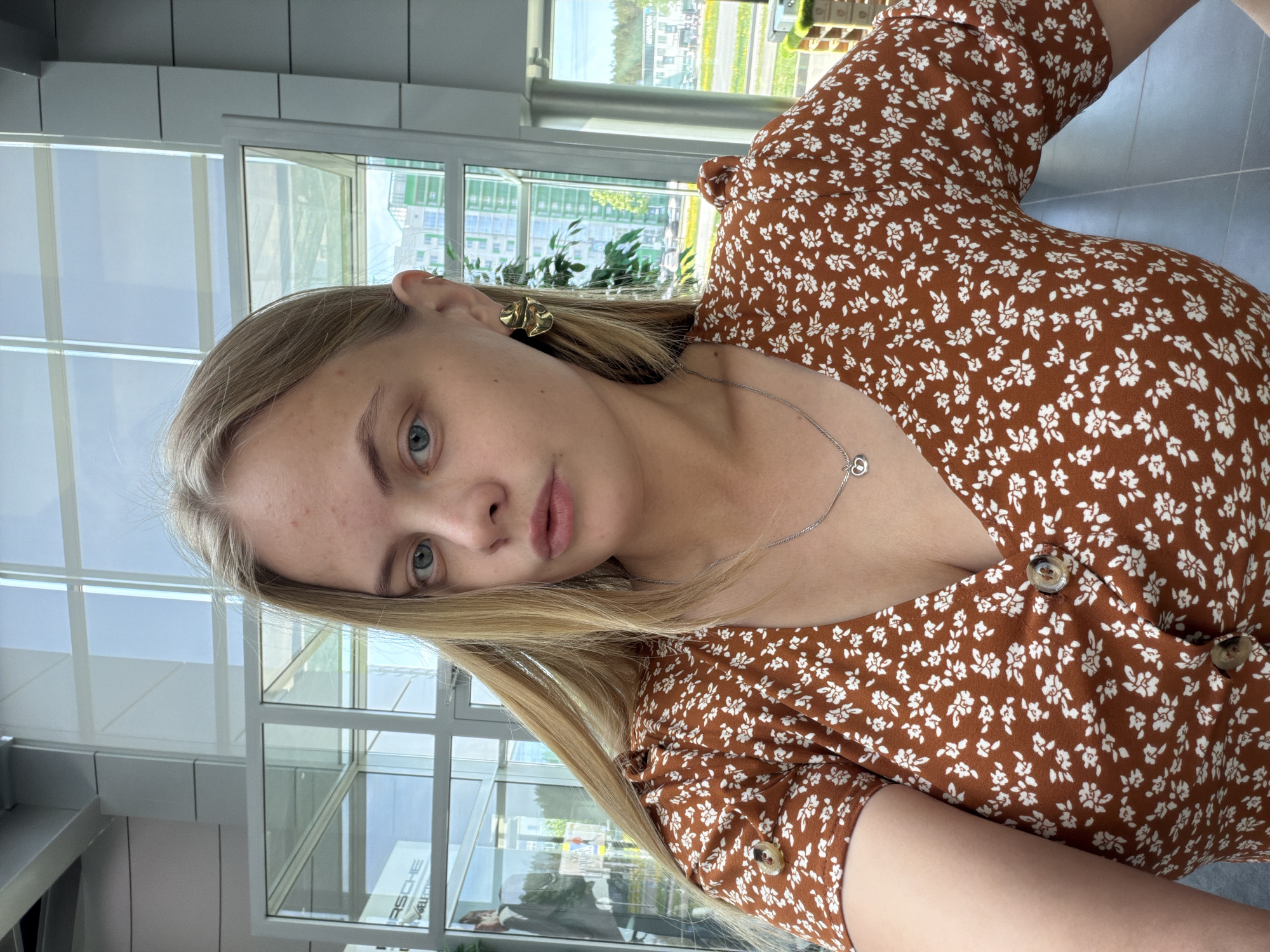 A young woman with light blonde hair and fair skin, wearing a brown dress with a white floral pattern, is the primary subject of this close-up photograph. She is facing the camera with a slight tilt of her head. She is wearing gold-colored earrings and a silver necklace with a heart-shaped pendant. The background features a bright, modern architectural space with large windows. Outside the windows, there's a cityscape with green spaces and buildings, suggesting a developed urban area. A sign with the word "Porsche" is visible through the window, indicating the potential presence of a car dealership or showroom in the vicinity. The image appears to be taken indoors, possibly in a building with a view of the city. It's not possible to confirm the specific location in the city of Izhevsk, Russia from the given image.