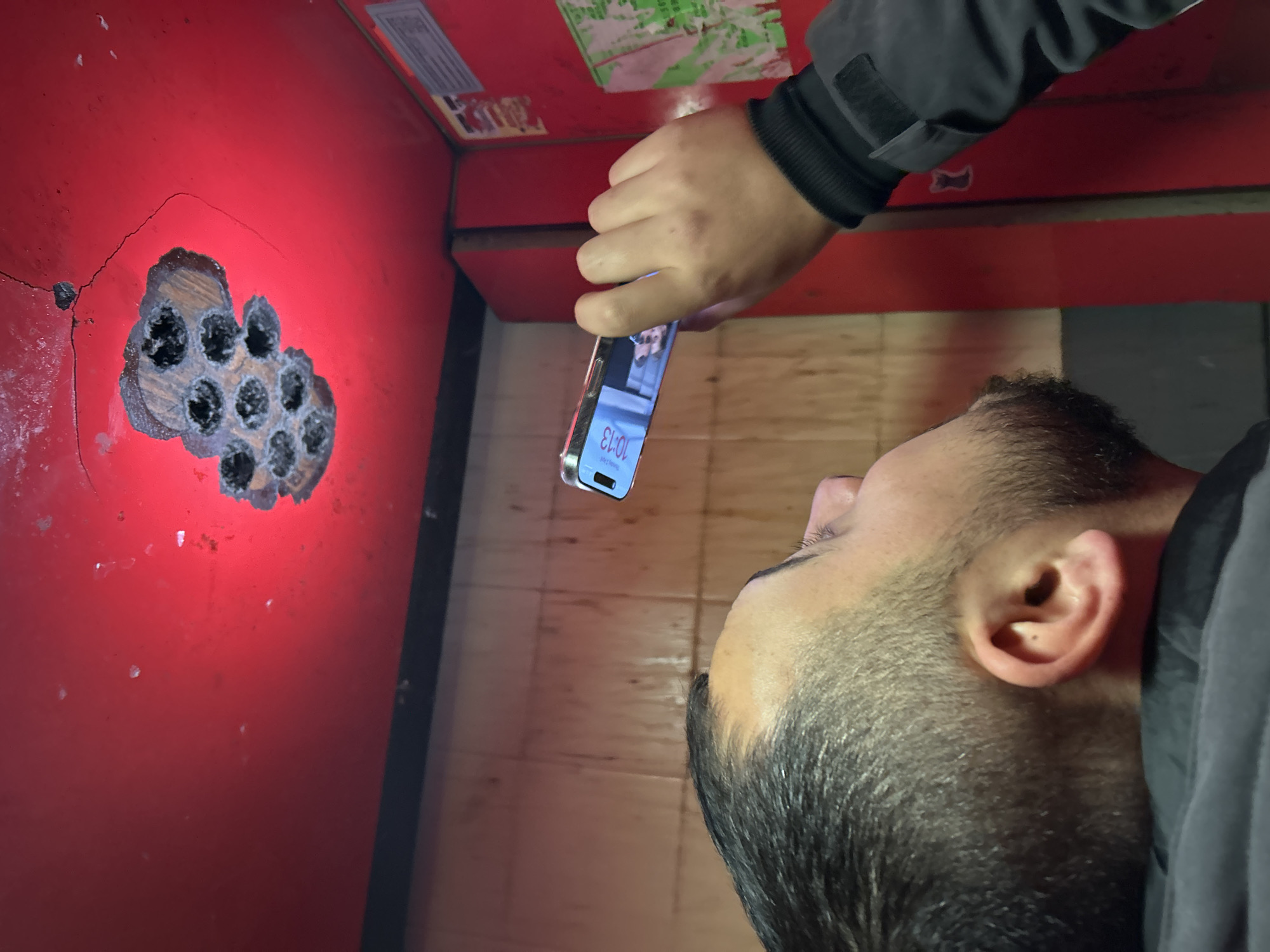 A man is holding a smartphone and looking at a red metal wall with a damaged area. The damaged area appears to have multiple small holes, possibly caused by drilling. The environment appears to be indoors, with tile flooring visible. The time on the smartphone screen reads 10:13. Sofia, Bulgaria is not discernable from this image.