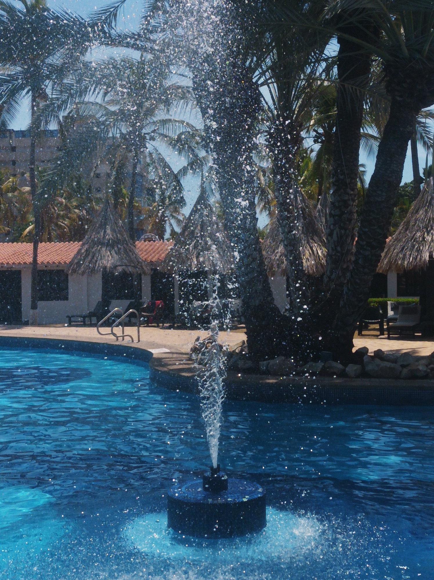 This vibrant photograph captures a resort swimming pool area in Porlamar, Venezuela, under bright daylight. The main subject is a water fountain situated in the foreground of the pool, actively spraying water upwards and outwards. Countless sparkling water droplets are suspended in the air, creating a dynamic and refreshing effect.
The pool itself features clear, bright blue water, with visible ripples and reflections on its surface. Beyond the pool, the setting is distinctly tropical, dominated by numerous tall palm trees with their fronds silhouetted against a clear, light blue sky. Behind the palm trees, various buildings are visible. To the left, a multi-story building with a light-colored facade can be seen through the foliage. Closer to the pool, there is a building with a distinctive red-tiled roof and several dark, possibly open-air, structures with conical thatched roofs, typical of a tropical resort.
The poolside area is paved and features lounge chairs, though no people are present. A silver handrail for entering the pool is also visible. The overall scene conveys a sense of a sunny, warm, and inviting vacation spot. No visible text is present in the image.
