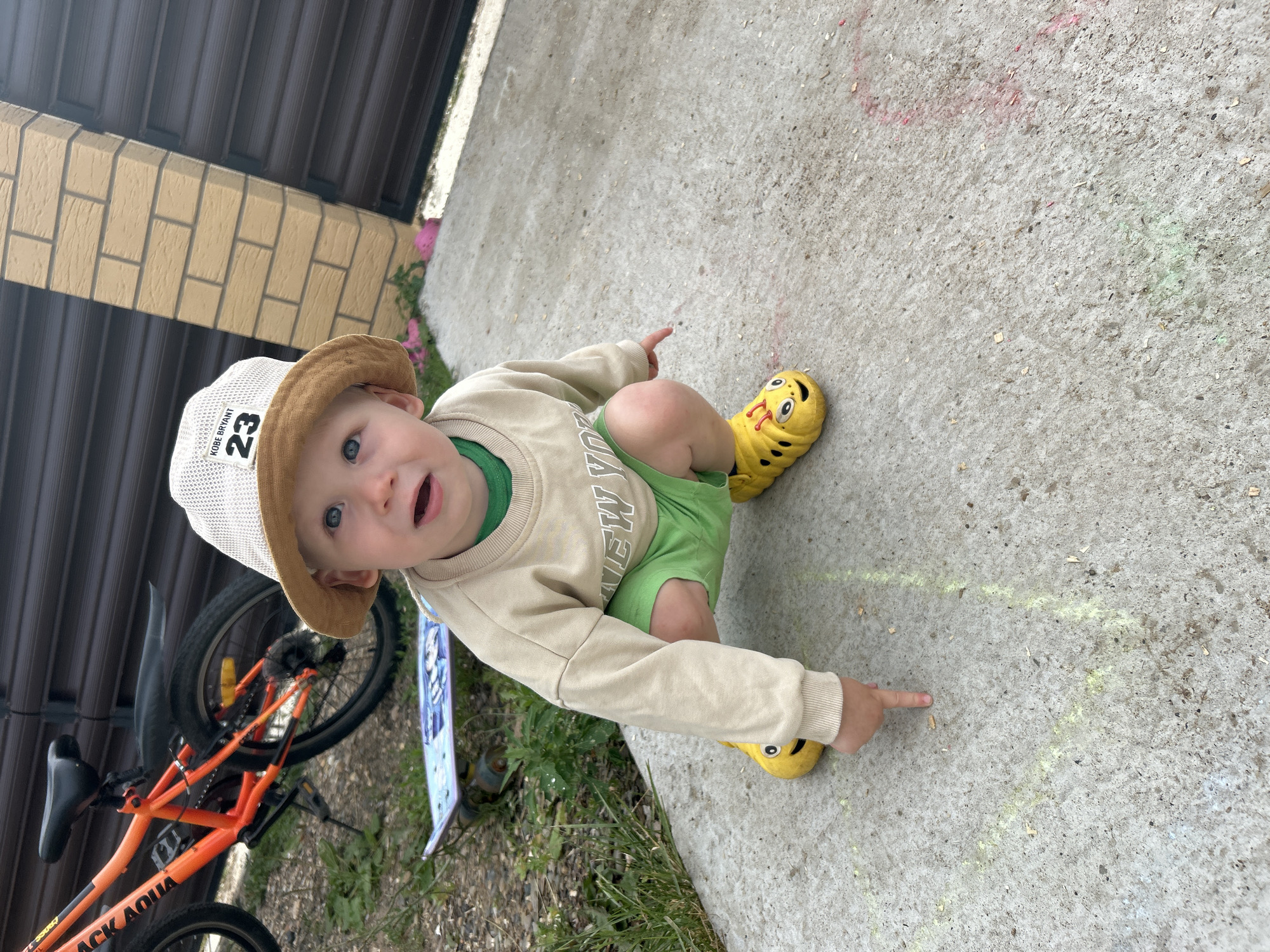 A young Caucasian child, with blonde hair and blue eyes, wearing a light brown bucket hat with a black inscription that reads "KOBE BRYANT 23", a tan sweatshirt over a green t-shirt, and bright green shorts, kneels on a concrete surface. The child wears yellow, bee-shaped clogs. The child is pointing a finger towards the ground, where chalk drawings are visible. In the background, an orange bicycle with a black fender is parked next to a dark brown wooden fence. A skateboard with a blue and white design lies on the grass to the left of the child. The scene appears to be outdoors, possibly in a residential area. The image does not provide specific geographical markers to confirm its location within the city of Chaikovsky, Russia.