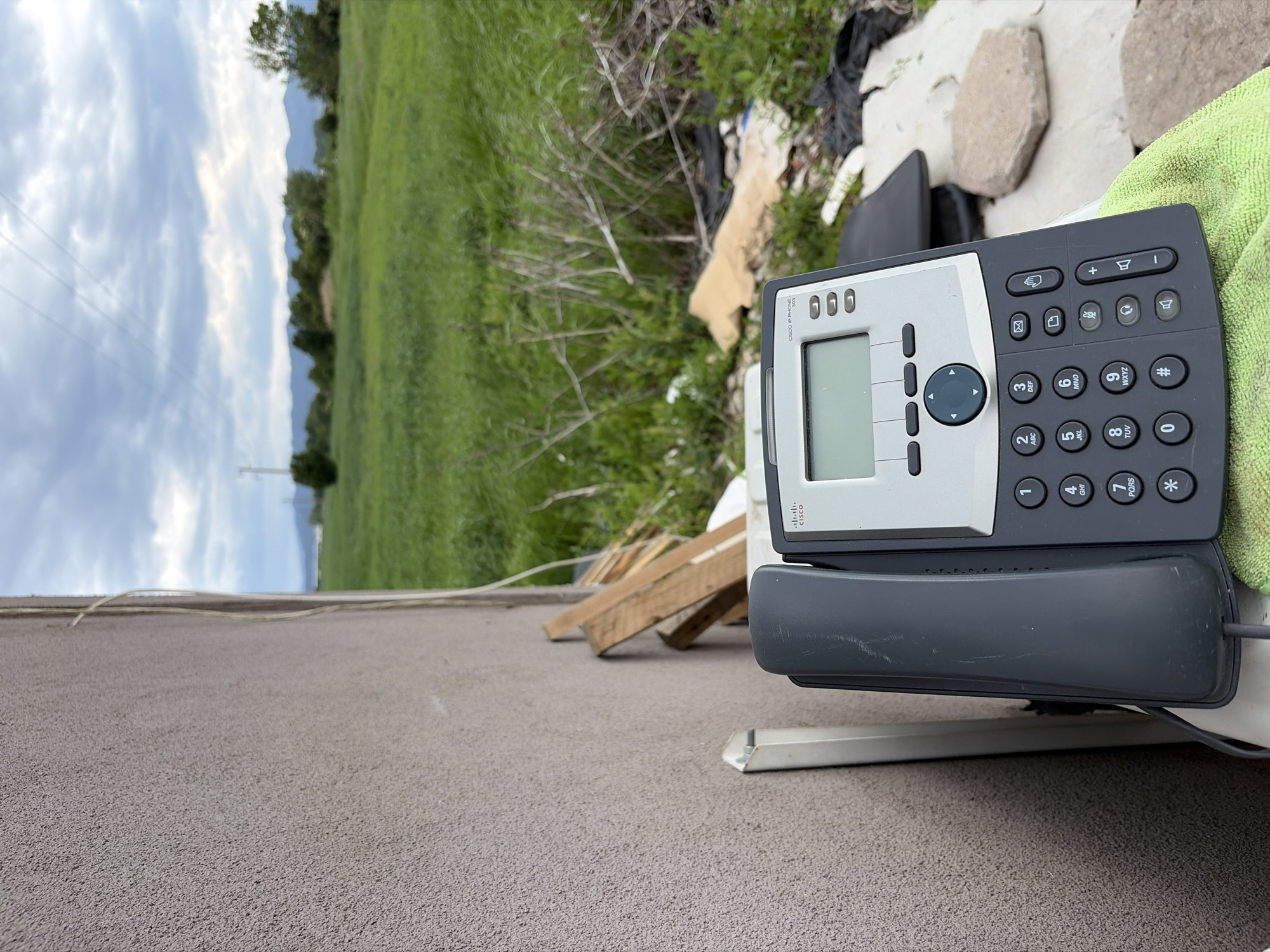A Cisco IP phone sits discarded on a gray surface, possibly asphalt, with a blurry green grassy area and overcast sky in the background. Debris such as broken wood, white fragments, and rocks are scattered near the phone. The location cannot be determined to be Sofia, Bulgaria with the given information.