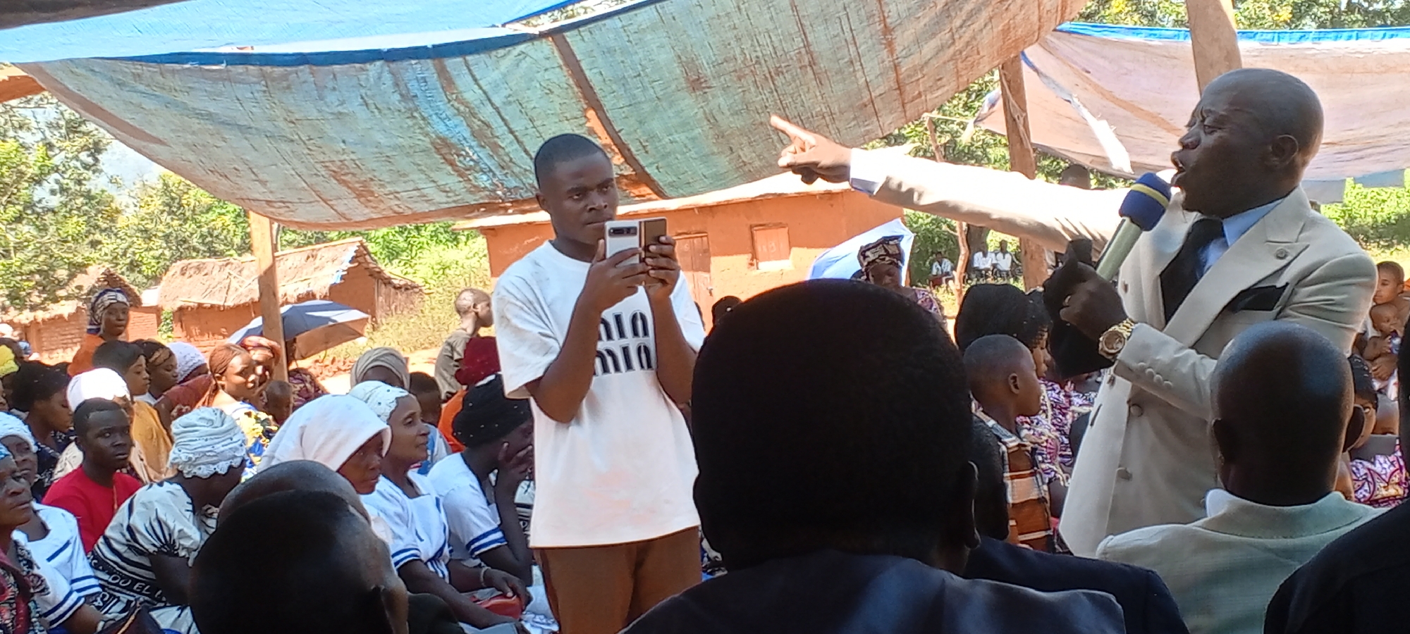 The image shows a gathering of people in what appears to be a rural area of Kalemie, Democratic Republic of the Congo. The setting is outdoors, possibly under a makeshift canopy of blue tarpaulin. Traditional houses with thatched roofs are visible in the background.
A man in a light-colored suit, holding a microphone, is speaking or preaching while gesturing with his right hand. He is wearing a watch on his left wrist. A younger man is standing nearby, holding up a smartphone, seemingly taking pictures or recording video.
Several individuals are seated, mostly women wearing traditional clothing and head coverings. The composition suggests a large crowd present. A few children are among the crowd.