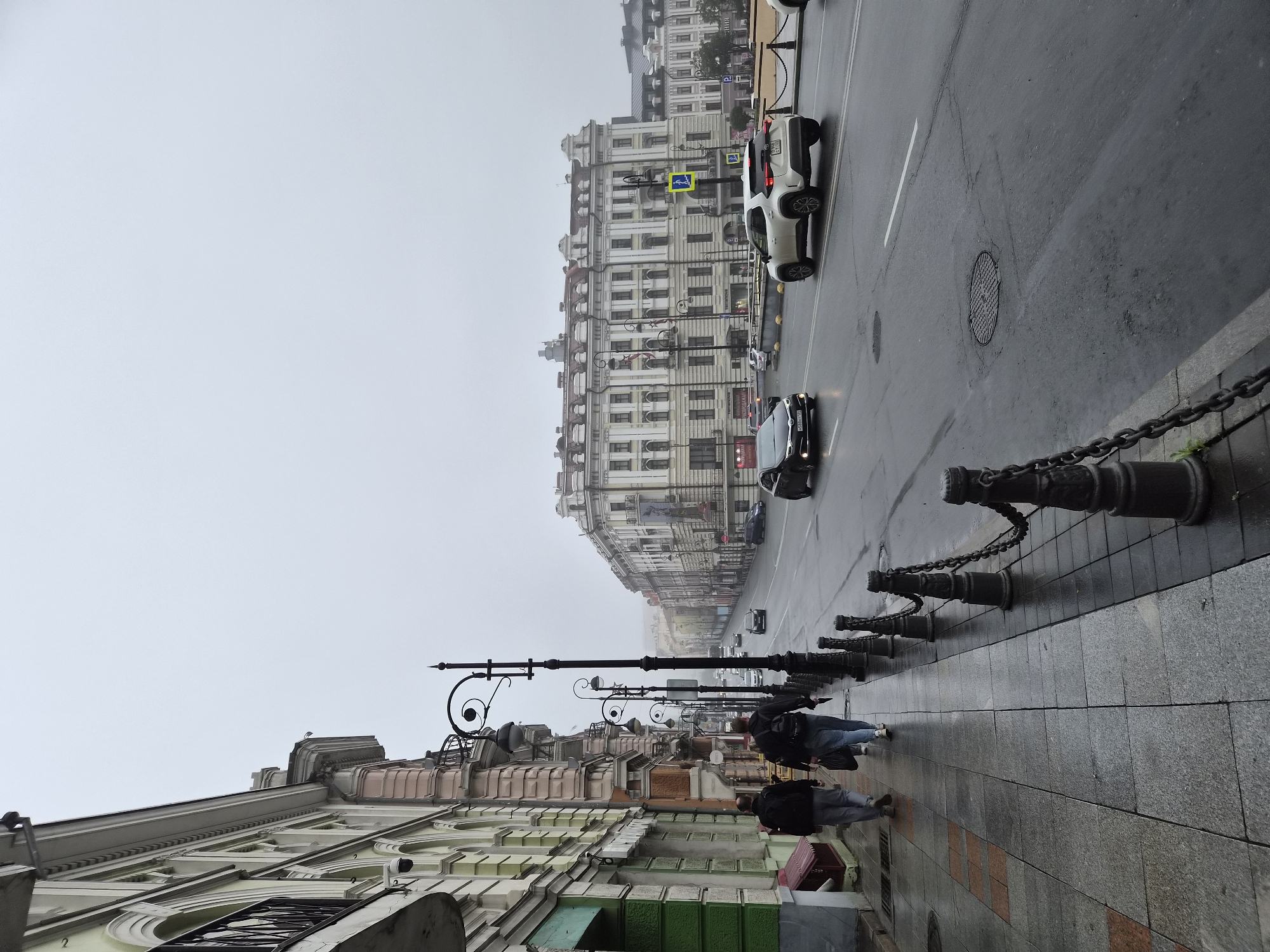 The image captures a cloudy day scene on a city street in Vladivostok, Russia. Two individuals are walking away from the camera on a wet sidewalk. The buildings lining the street exhibit classic architectural styles with ornate facades and balconies. A streetlamp with a decorative spiral element stands prominently on the sidewalk. Cars are driving on the wet asphalt road. The overall atmosphere suggests a typical urban environment on an overcast day.