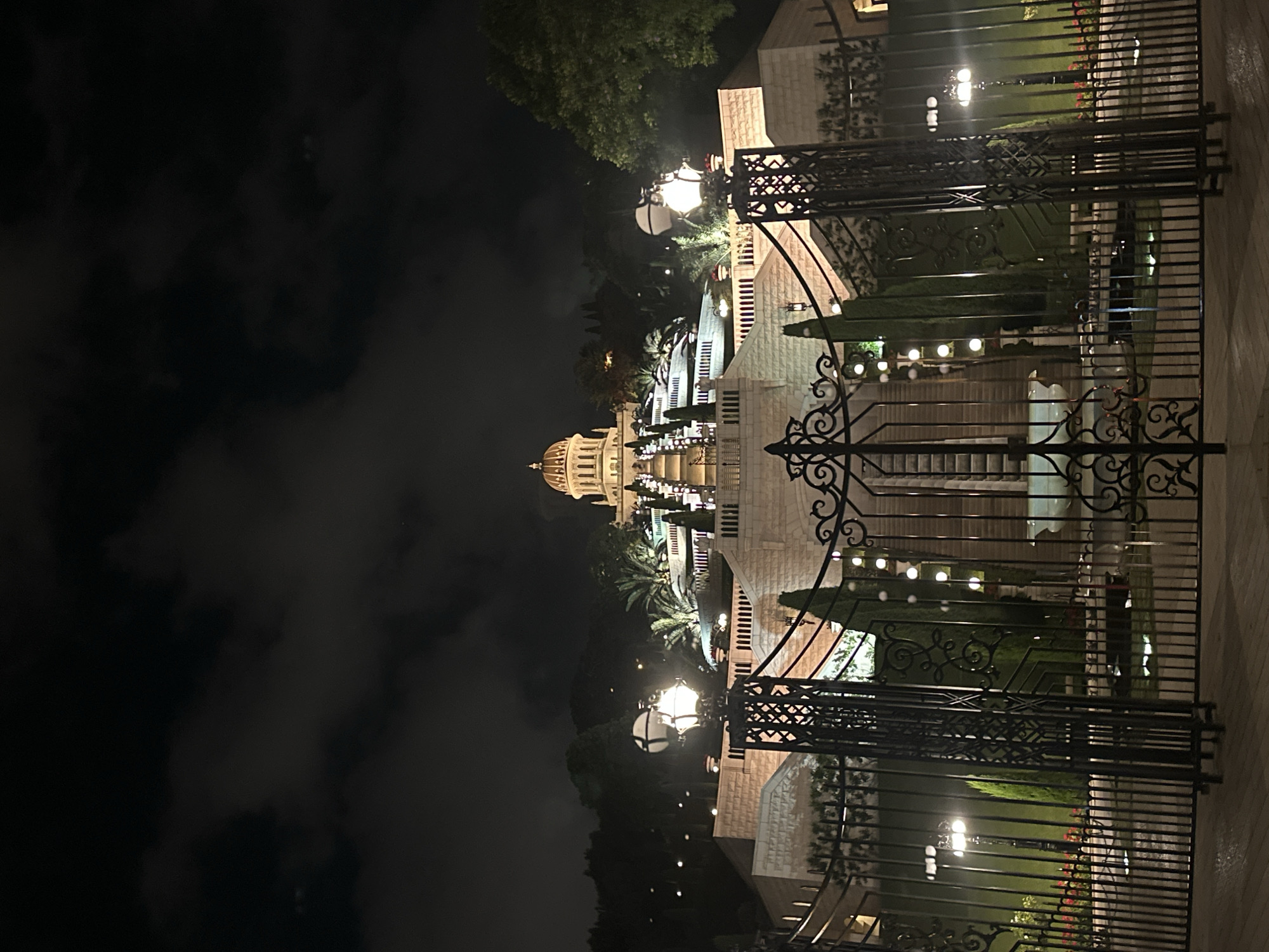 The image depicts the Baha'i World Centre on Mount Carmel in Haifa, Israel, at night. The iconic golden-domed building is illuminated and visible at the top of a terraced staircase. The terraces are adorned with manicured greenery, palm trees, and decorative lighting. In the foreground, a large, ornate black metal gate stands closed, with decorative finials and vertical bars. Two lampposts with globe-shaped lights are visible just beyond the gate. The overall scene is dramatic, with dark, cloudy skies contrasting with the bright illumination of the Baha'i complex. No people are visible in the image. The environment appears to be a garden or landscaped area with stone pathways and walls.