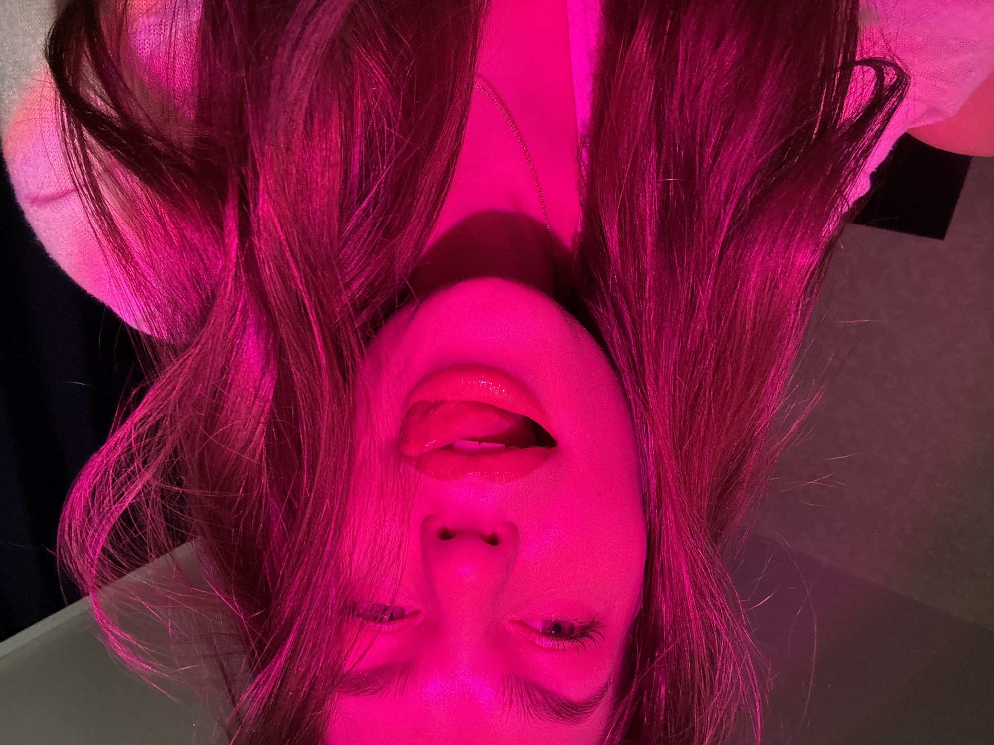 A portrait of a woman illuminated by pink lighting, taken from an upside-down perspective. The woman's mouth is open with her tongue extended. She has long hair. A thin necklace is visible on her neck. The location is unknown, and there is no information about it being taken in the city of Chaikovsky, Russia. The environment is partially visible with sections of what could be a table and draped fabric in the background.