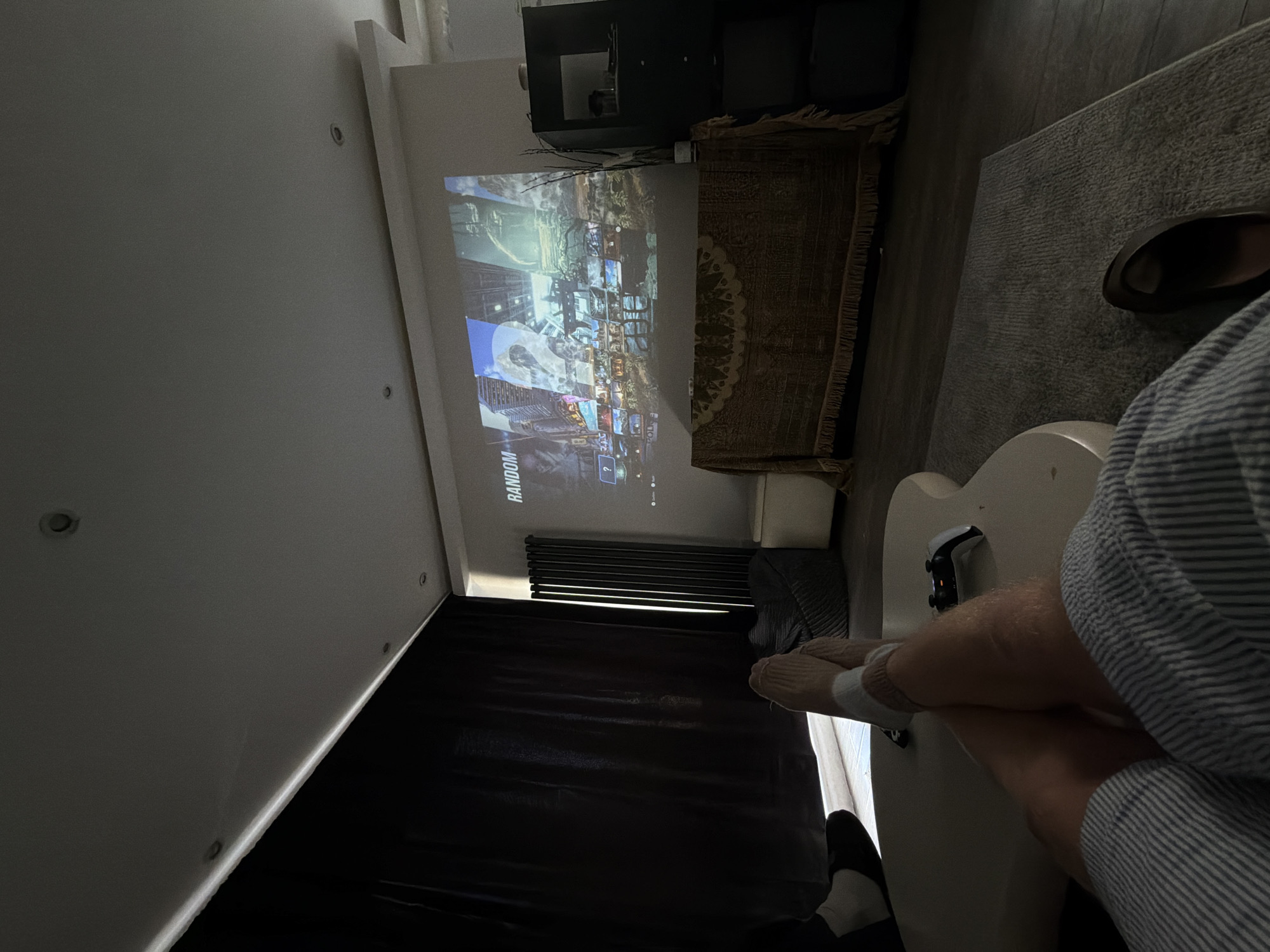 The image captures a first-person perspective of a relaxed individual engaging in a video game session in an indoor setting. The main subject is the person's lower body, with legs propped up, clad in socks and a striped shirt sleeve. A white gaming controller, resembling a PlayStation 5 DualSense, is visible on the surface where the person's feet are resting.
The focal point of the room is a large image projected onto a light-colored wall, displaying what appears to be a video game, possibly "Call of Duty," judging by the fragmented tactical imagery and the visible text "RANDOM" and a question mark icon within the game interface. The projector itself is situated on a dark shelf or unit above the projected image.
The room is dimly lit, suggesting it's either evening/night or that dark curtains, visible in the lower left, have been drawn to optimize projection viewing. Recessed lighting fixtures are embedded in the white ceiling. The floor features a textured grey rug, and a patterned, fringed rug lies beneath the projector setup. A pair of dark slippers or sandals are casually placed on the rug near the person's feet. The overall ambiance is one of comfortable leisure and entertainment. The location is an indoor residential space, potentially a living room.