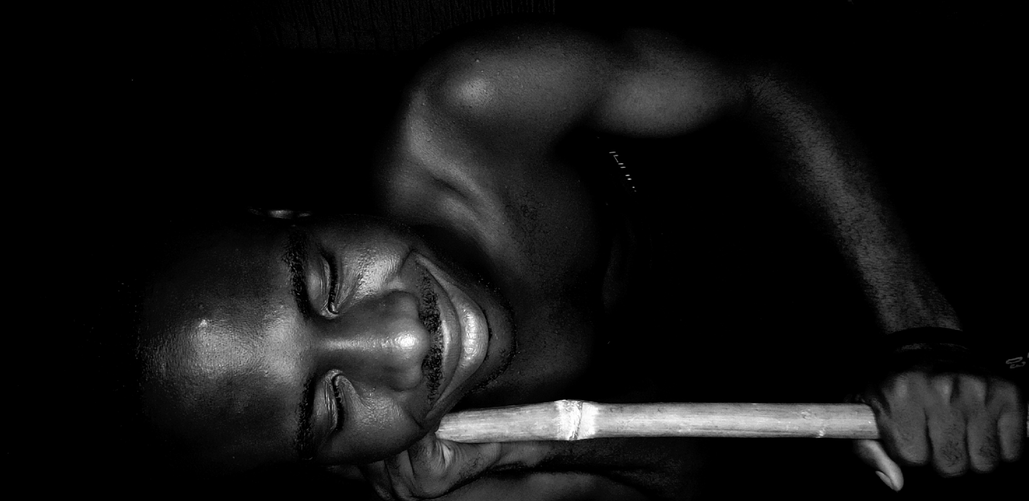 A man with dark skin is lying down, his eyes closed, and holding a light-colored, cylindrical object in his hand. The scene is dimly lit, with the subject’s face and upper body visible against a dark background. A partial view of a shoulder, an arm, and a hand gripping the cylinder is discernible. The location is Narayi, Nigeria.