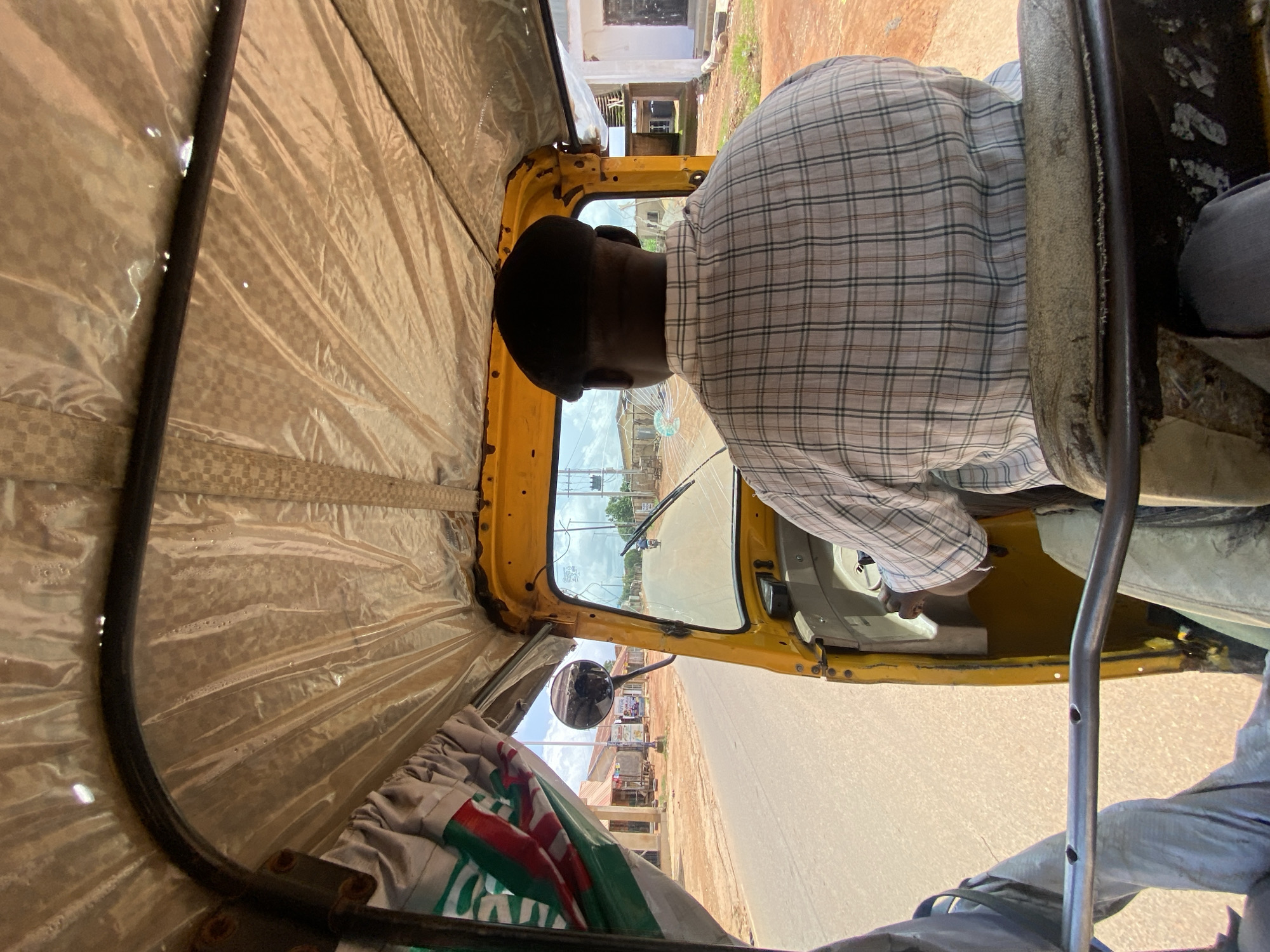 The image shows the interior of a yellow auto-rickshaw traveling on a road in Gura Top, Nigeria. The driver, a Black man wearing a dark cap and a plaid shirt, is seated in the driver's seat and facing forward, controlling the vehicle. Through the windshield, a paved road is visible, flanked by buildings and sparse vegetation. A single motorcycle rider is seen in the distance. The ceiling of the auto-rickshaw is covered with a tan, textured material, and a green, white, and red striped curtain is partially visible on the left side. The overall lighting suggests it is daytime.