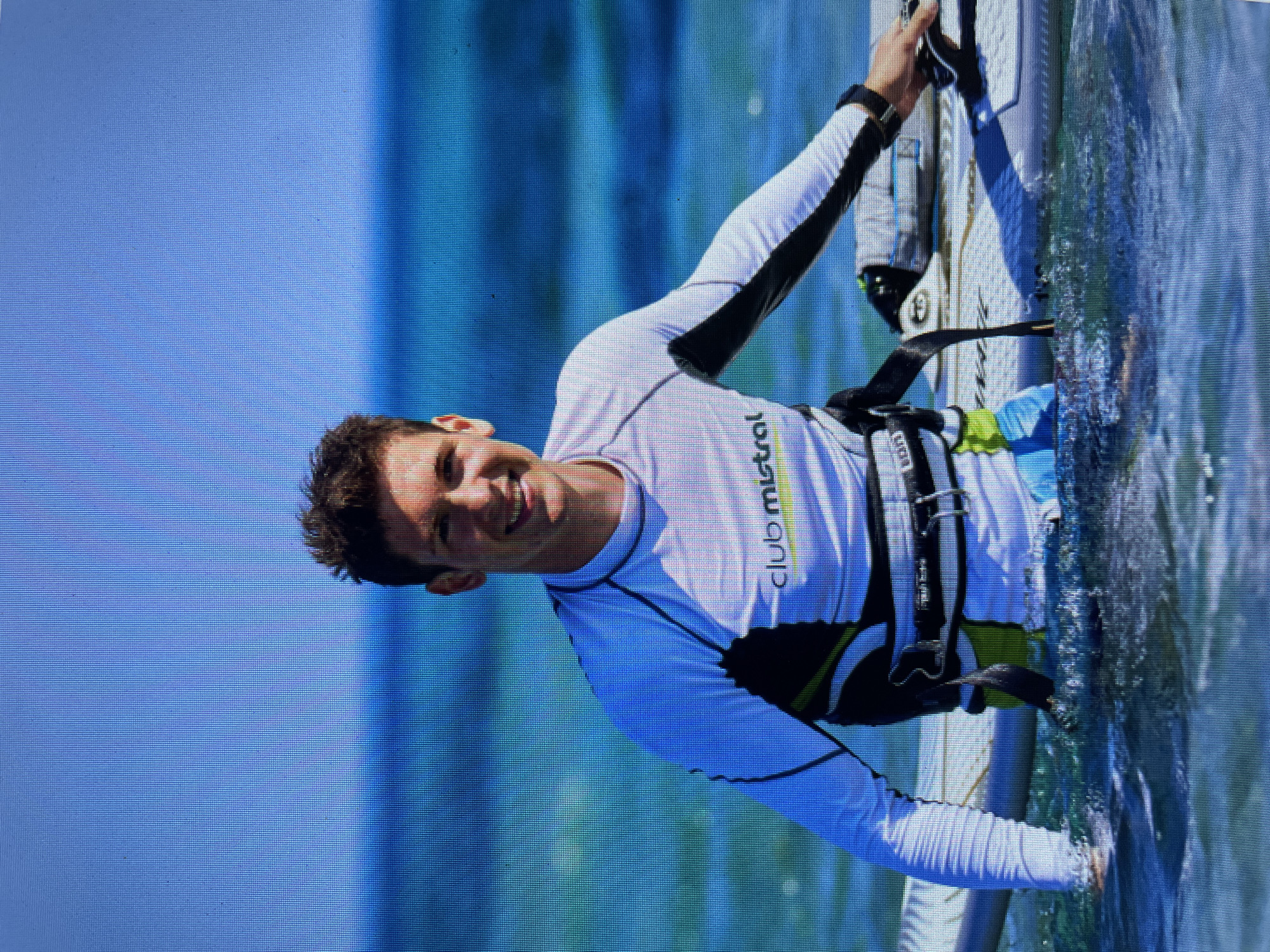 A smiling man is windsurfing in Bacong, Philippines. He's wearing a white long-sleeved shirt with black accents and a black harness. He is holding onto the windsurfing board, which is partially submerged in the water. The water is turquoise, and the sky is light blue. The Club Mistral logo is visible on his shirt.