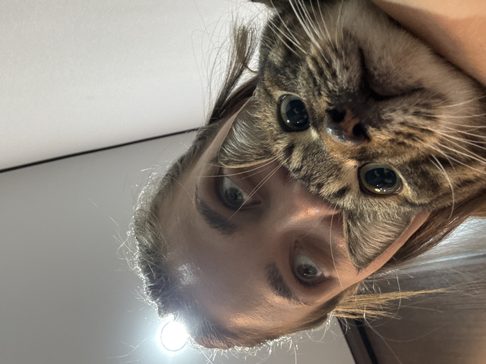 A close-up, upside-down image shows a woman's face next to a tabby cat's face. The woman has light skin, brown eyebrows, and light-colored eyes. The cat has brown and tan fur with large, round, black eyes. They are both looking at the camera. The background is a blurry ceiling. There is no indication that this is in Chernushka, Russia.