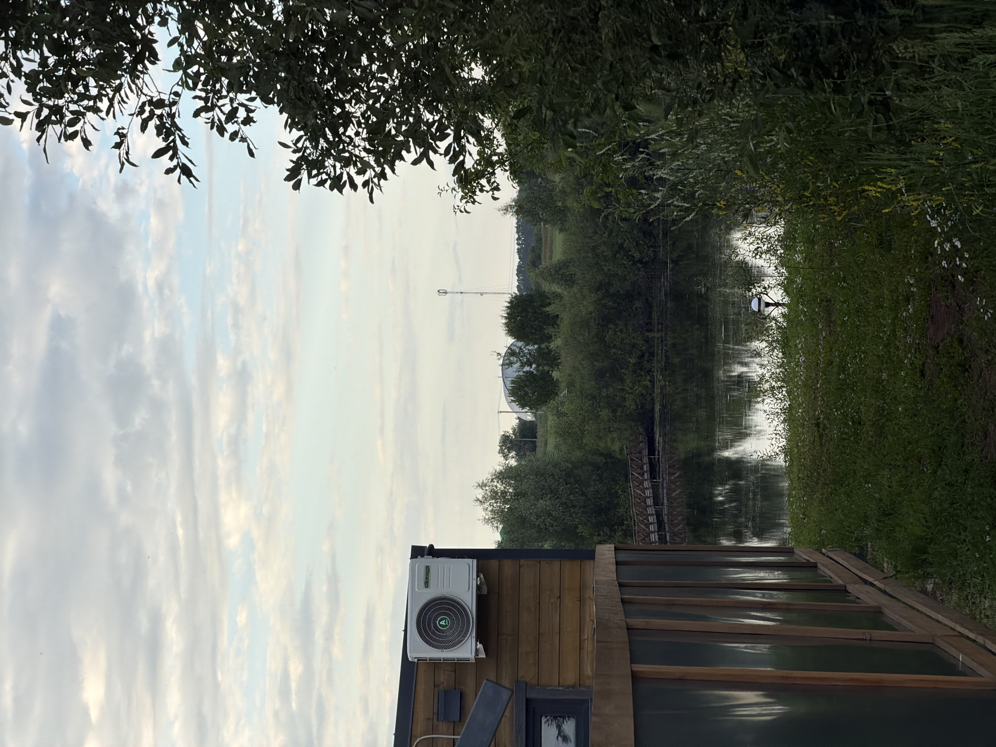 This image captures a serene waterside scene in Bol'shaya Ven'ya, Russia. The primary subjects are a body of water, likely a river or canal, and a small wooden structure with an attached air conditioning unit. Lush green trees and foliage line the banks of the water, creating a natural and somewhat wild atmosphere. In the distance, a tall radio tower and some rounded structures are visible, suggesting a blend of nature and human development. The sky is overcast with soft, diffused light, indicating it might be early morning or late afternoon. There are no people visible in the image, and no specific activity or event appears to be taking place, lending a sense of stillness to the scene. The wooden structure, with its visible planks and glass panels, suggests it might be a small dwelling or a shed. The overall mood is tranquil and peaceful, with the natural elements dominating the composition.