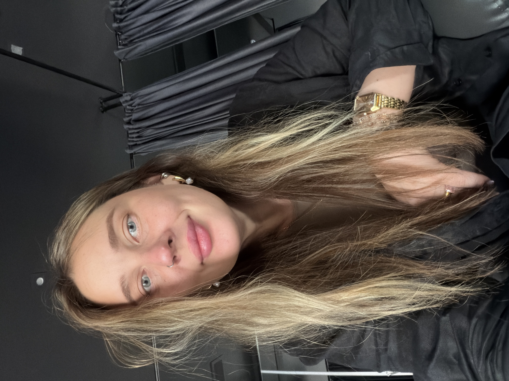A woman with long blonde and brown hair, light skin, and blue eyes is shown in a medium close-up selfie. She wears a black shirt, a gold watch, and multiple bracelets on her left wrist, as well as earrings and a nose ring. In the background are dark gray drapes and a dark gray wall. The setting appears to be indoors. There is no specific information available to determine if the location is in Moscow, Russia.