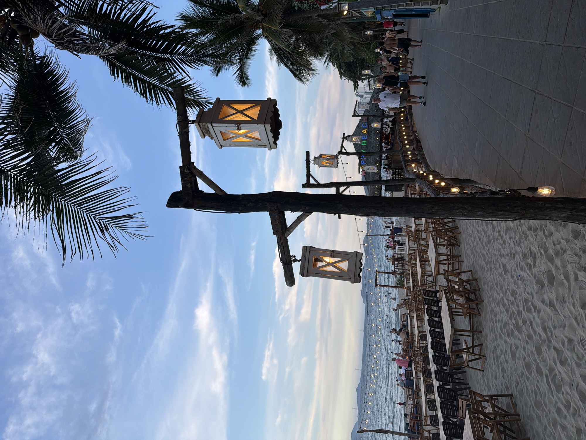 The image depicts a beachside scene in Nha Trang Ward, Vietnam. In the foreground, a sandy beach is covered with numerous wooden tables and chairs, suggesting a dining area. Palm trees with their fronds arch over the scene. Two wooden posts with decorative lanterns hang from them. The lanterns have a weathered, wooden exterior with glass panes, illuminating a warm, orange light. Along a paved walkway, several people are walking. The walkway is lined with a wooden railing decorated with string lights. Further in the background, the ocean is visible with many people swimming and enjoying the water. A few buildings and a sign that reads "HAPPY BEACH" are also visible in the distance. The sky is partly cloudy, with a soft blue hue, indicating it is either early morning or late afternoon.