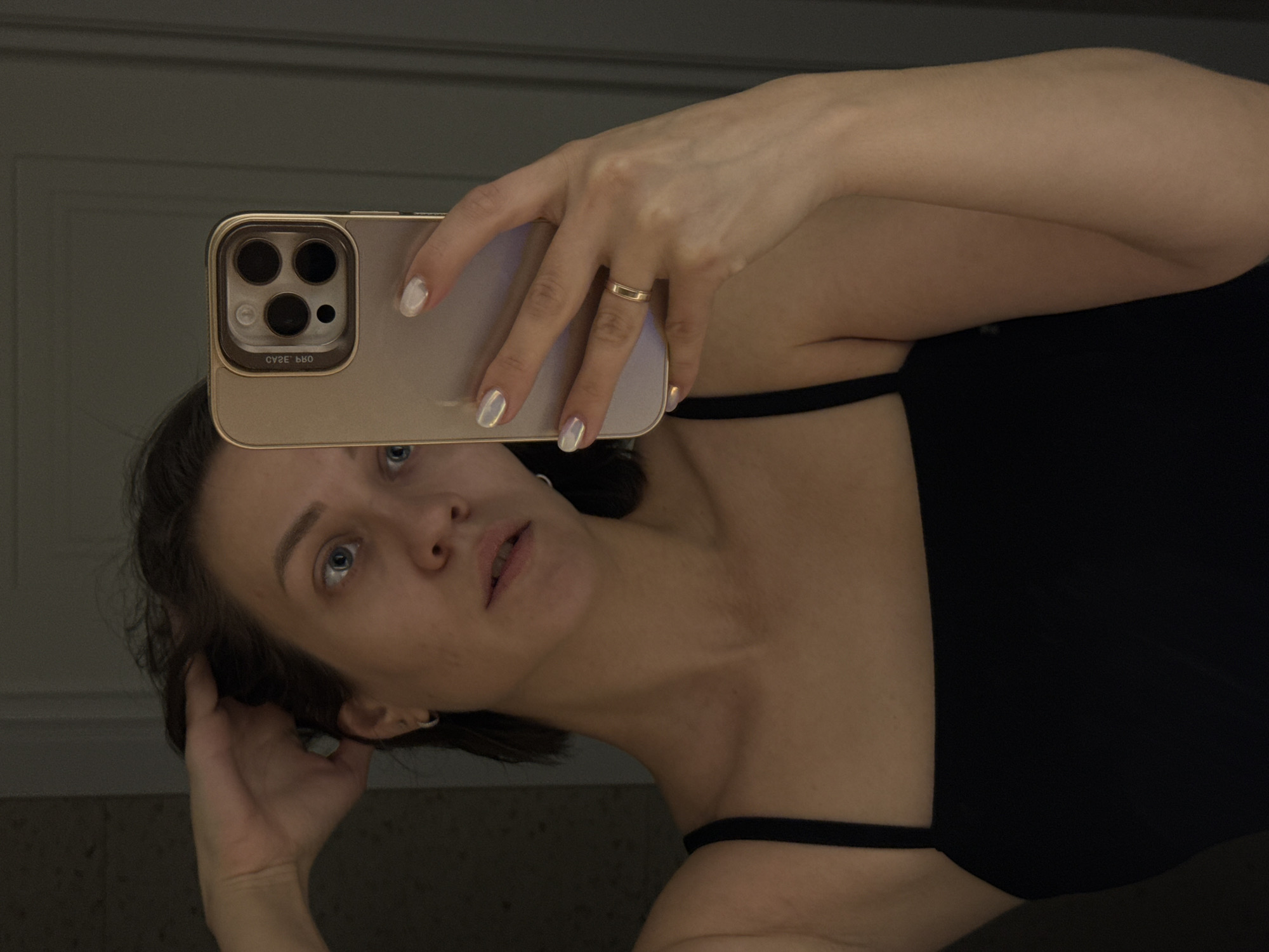 The image is a mirror selfie depicting a young adult woman indoors. She holds a gold-colored smartphone, featuring a prominent camera module with three lenses and a flash, in her right hand to capture her reflection. Her head is tilted, and her blue eyes are directed upwards, possibly at the phone screen or the mirror itself. Her left hand is casually raised to her dark hair, near her ear.
She is wearing a dark top with thin straps, likely a camisole or tank top. A simple, thin gold-colored band ring is worn on the ring finger of her right hand. Her fingernails are painted with a subtle, iridescent light-colored polish. The background behind her reflection is dark and indistinct, suggesting an interior setting like a room or bathroom with low lighting.
Visible text on the phone case's camera module is inverted, reading "CVEE bBO," which appears to be a reversed brand name such as "CASE LAB." The lighting in the scene is dim, creating a subdued atmosphere. The woman's expression is neutral and focused on the act of taking the self-portrait.