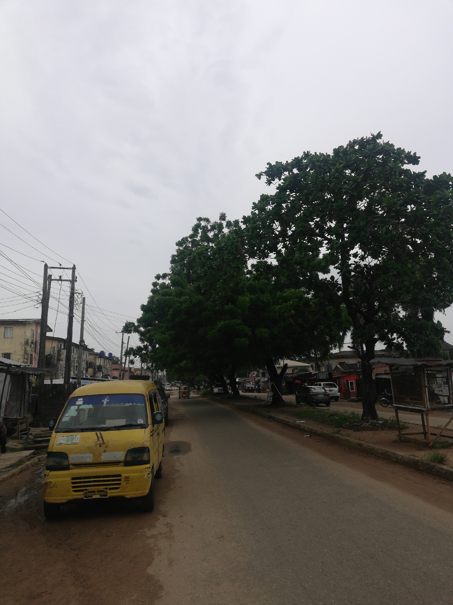 The image depicts a street scene in Lagos, Nigeria, under an overcast sky. A yellow minibus-style taxi is parked on the left side of the road, appearing worn with some visible damage. Utility poles and wires line the left side of the street alongside older buildings. The street is asphalt and appears damp in places. On the right side of the road, large trees cast shade, and there are a few parked vehicles visible further down the street, along with a small building and makeshift structures. The overall impression is that of a typical urban street in Lagos.