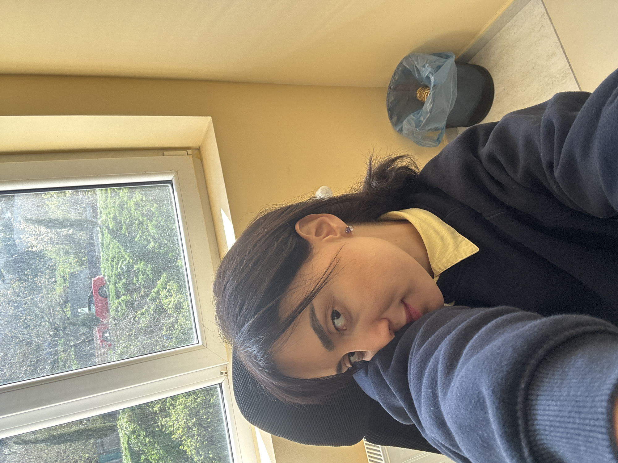A selfie taken indoors, likely during the day, shows a person with dark hair and light skin leaning against a black mesh office chair. The person wears a dark blue sweater over a yellow collared shirt. They are positioned in front of a window, through which trees are visible, along with a glimpse of a red structure or object. The room has pale yellow walls. A wastebasket with a blue plastic liner and visible contents is partially visible in the upper right corner. This image was most likely taken in an office building or residence hall in the city of Tbilisi, Georgia.