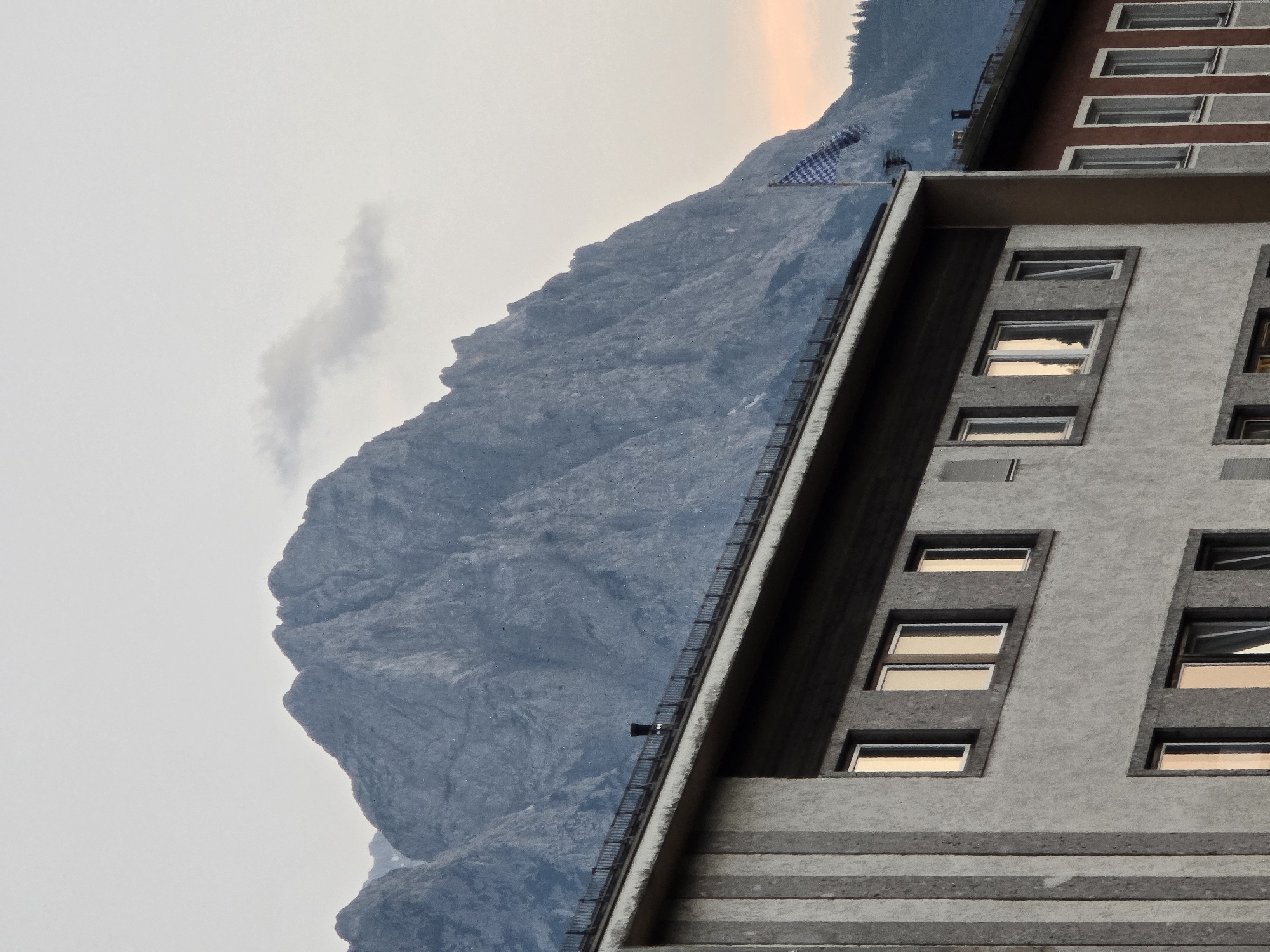 This image captures a dramatic mountain landscape behind a modern building. The mountains, with their rugged, textured surfaces, fill the background and a significant portion of the frame. A thin wisp of cloud is visible against the pale sky. A flag with a blue and white checkered pattern is planted on a pole on the mountain slope, near the summit.
In the foreground, a multi-story building with a grey, textured facade is prominent. The building features numerous rectangular windows, many of which reflect the warm, golden light of either sunrise or sunset. The architecture appears contemporary, with clean lines and a geometric arrangement of windows. Another building, partially visible to the right, has a reddish-brown facade with a similar window pattern.
The image conveys a sense of elevation and grandeur, with the imposing mountains juxtaposed against human-made structures. The soft light suggests it is either early morning or late evening. The overall mood is serene and somewhat awe-inspiring, highlighting the interaction between nature and architecture. No visible text is present in the image. The location context provided is Garmisch-Partenkirchen, Germany, which aligns with the mountainous terrain.