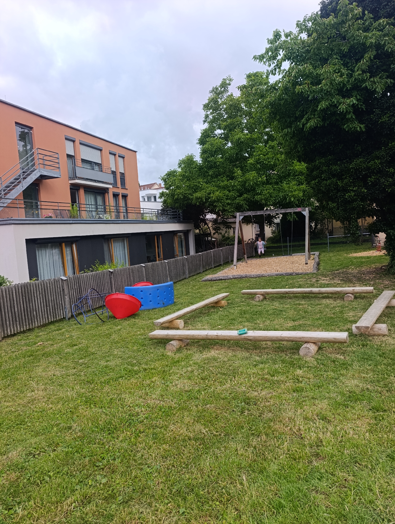 This image depicts an outdoor children's play area located in Pfullingen, Germany. A modern, orange-facade apartment building is visible in the background, featuring balconies and an external staircase. Adjacent to the building, a wooden fence encloses a grassy area containing playground equipment.
The playground includes a swing set with a child on one of the swings, a blue climbing structure, and a red, dome-shaped object that appears to be part of a larger play apparatus. Several low, wooden beam structures are arranged on the grass, possibly serving as balance beams or stepping stones. The ground is covered in green grass, with some patches of sparser vegetation. Trees with dense green foliage are situated behind the playground equipment and the building, suggesting a residential or park-like setting within the city. The sky is overcast.