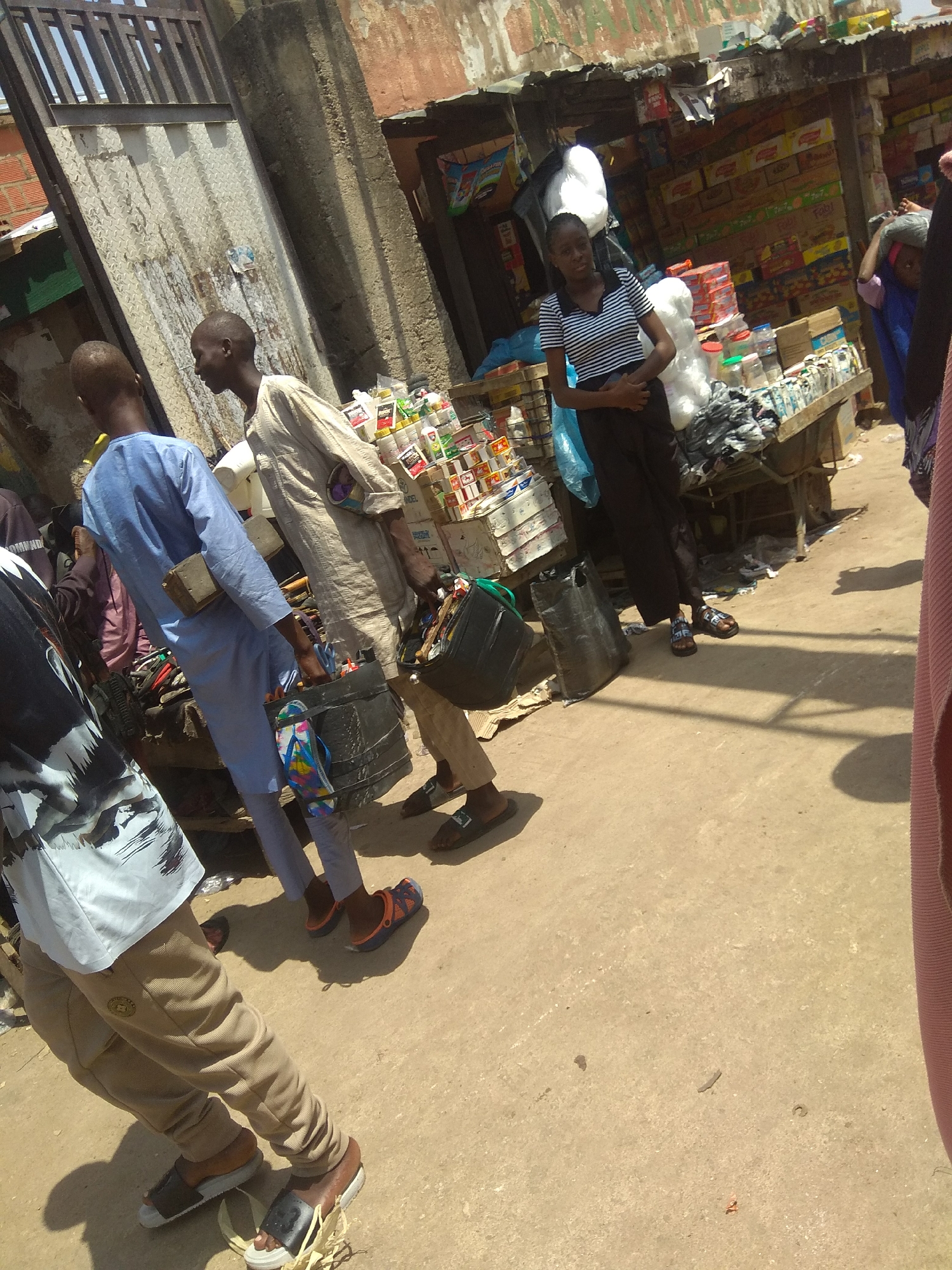 The image depicts a bustling outdoor market scene in Jalingo, Nigeria. In the foreground, several individuals are visible, engaged in various activities. One person, wearing a light blue garment, carries a black bucket containing a pair of colorful flip-flops, and a wooden object is strapped to their back. To their left, another person in a light-colored robe is also seen carrying a similar black bucket. In the middle ground, a woman in a striped t-shirt and dark trousers stands looking towards the camera, positioned near a stall filled with assorted goods. Further back, stacks of boxes and products are arranged on tables and shelves, suggesting a marketplace environment. The ground is dusty and uneven, characteristic of an outdoor market. The overall atmosphere appears active and commercial, with multiple people navigating the crowded space.