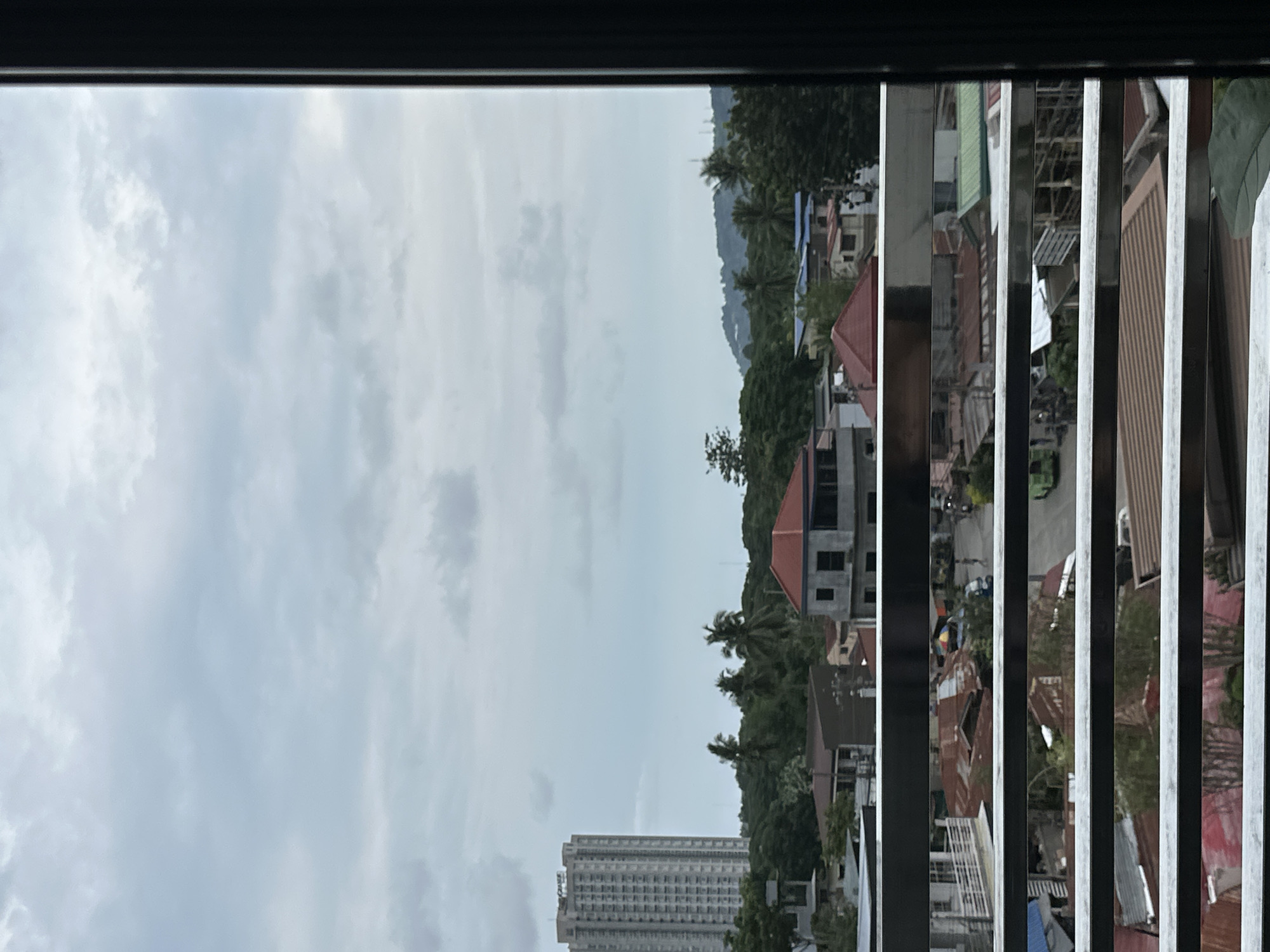 The image shows a view from a high vantage point, likely a window, overlooking a cityscape in Davao City, Philippines. The sky is overcast with grey clouds. The landscape includes buildings of varying heights, ranging from low residential structures with corrugated iron roofs to a taller, modern high-rise building in the distance. Lush green vegetation, including trees and possibly forested hills, is interspersed among the buildings. A metal window frame or bars partially obscure the view, creating a framed perspective of the urban scene.