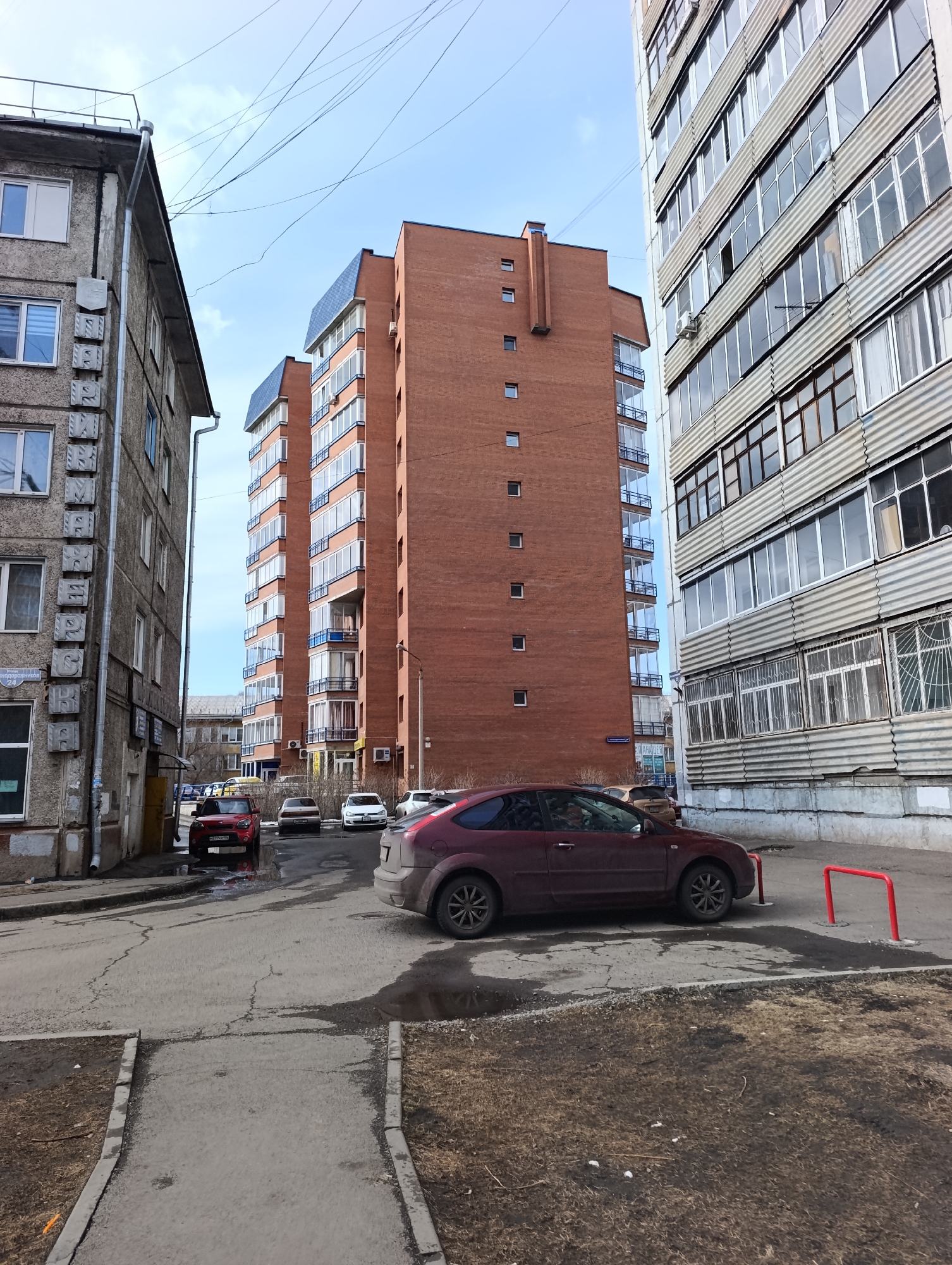 This full-shot, street-level photograph depicts an urban residential area in Krasnoyarsk, Russia, under a bright, partly cloudy sky. The scene is framed by three distinct apartment buildings. On the left, an older, grey concrete-paneled building with visible drainage pipes features a vertical Cyrillic sign clearly reading "ПРОСВЕЩЕНСКАЯ 24" (PROSVESHCHENSKAYA 24), indicating a street address. In the center-background stands a taller, modern red-brick building with numerous balconies showcasing blue glass railings and dark-sloped roofs. To the right, another multi-story grey building, similar in style to the one on the left, displays rows of windows, some with visible air conditioning units.
The paved ground in the foreground and midground shows signs of wear with cracks and some wet patches or puddles. Several cars are parked along the street or courtyard. A maroon hatchback is prominently positioned on the right, next to a red U-shaped metal parking barrier. Further back, a red SUV is parked near a puddle, its license plate "О 277 РН 190" (Moscow region) visible. Other parked vehicles, including a white sedan, are visible in the background. The ground cover consists of dry dirt and dormant grass, suggesting early spring or late autumn. Overhead, numerous utility wires crisscross against the sky. No people are visible in the frame, portraying a quiet, everyday urban scene.