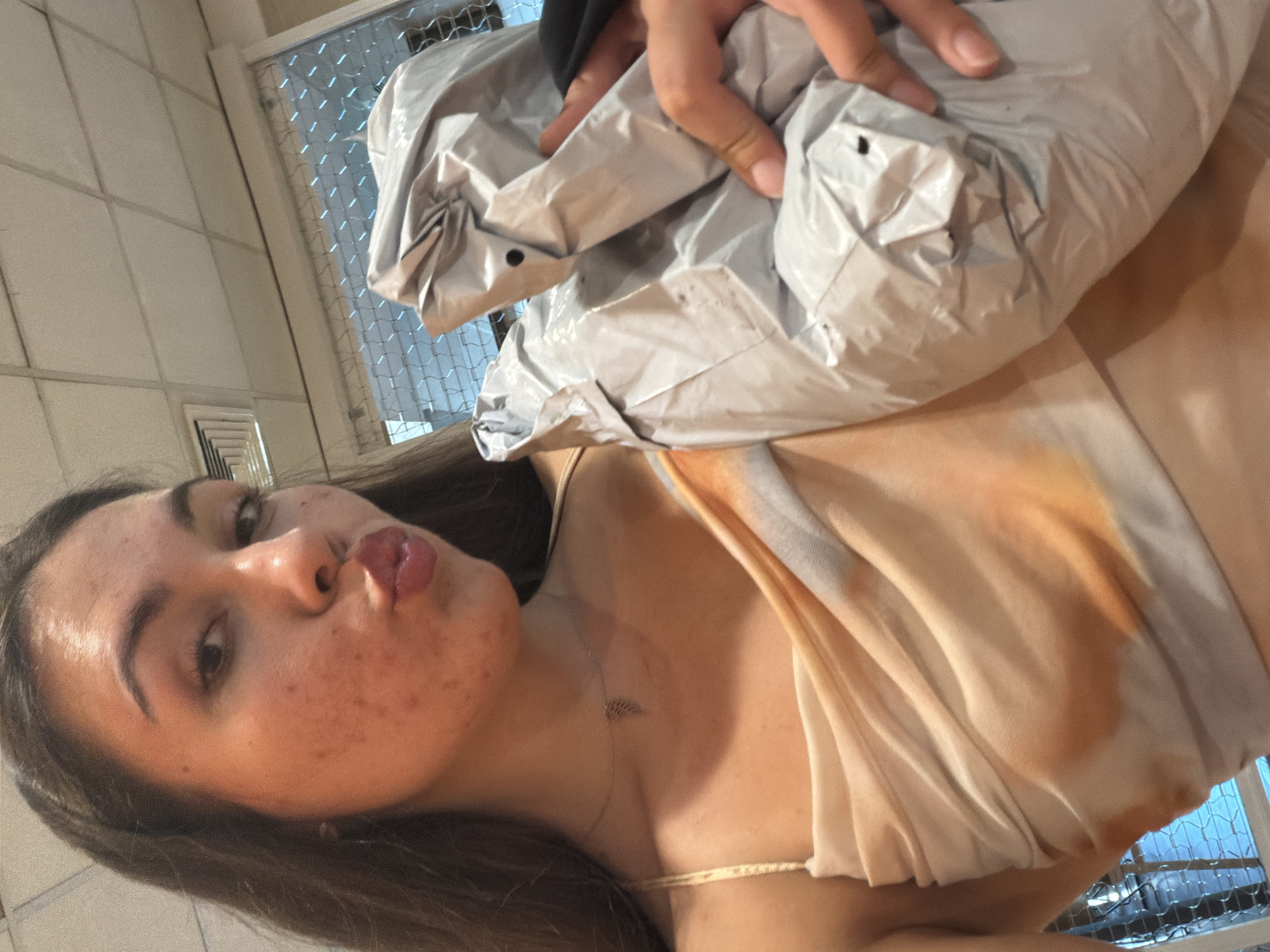 A young woman with dark hair, wearing a light-colored, draped top with an orange tie-dye pattern, is holding a crinkled silver package wrapped in plastic. She is puckering her lips and looking at the camera. She is wearing a thin silver necklace with a leaf-shaped pendant. The background appears to be an indoor setting with fluorescent lighting on a tiled ceiling and a window with a wire mesh security screen. This image was taken in Varna, Bulgaria.