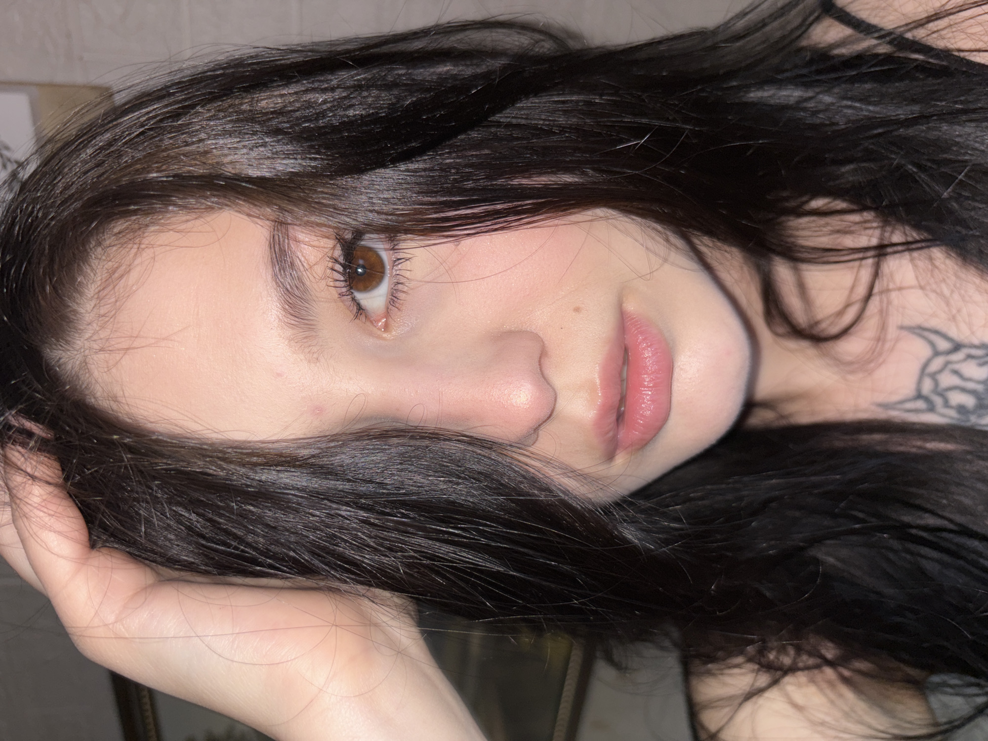 A close-up portrait of a fair-skinned person with dark brown hair partially obscuring the face. The person has brown eyes, noticeable eyebrows, and a mole on the cheek. Their lips are pink and appear glossy. A hand is visible holding the hair. A tattoo can be seen on the upper chest/shoulder area. The background is indistinct. There is no indication of Фоки, Russia, or any other geographical location.