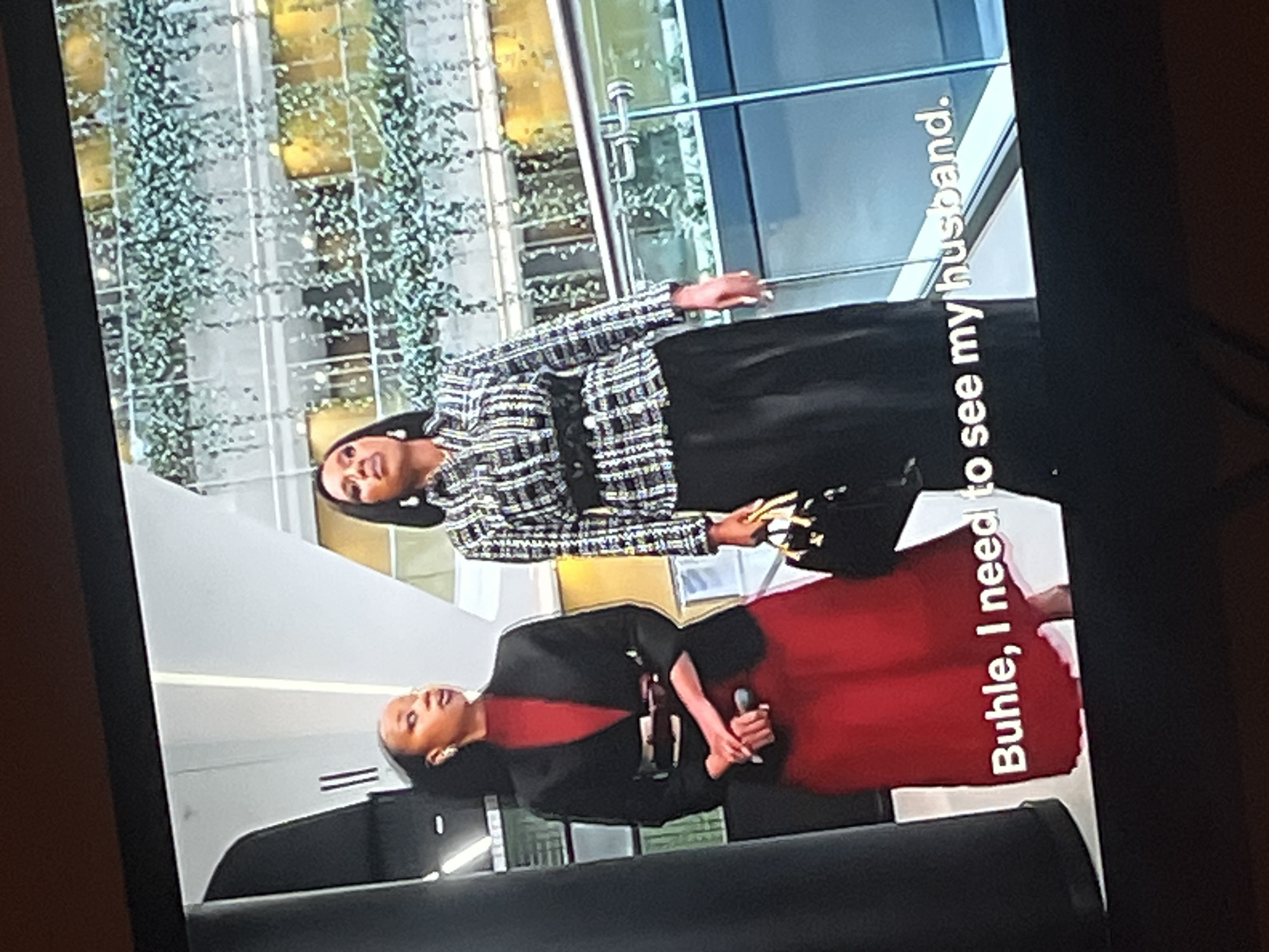 The image shows two women standing on a balcony in an indoor atrium. The woman on the left is wearing a red dress and a black blazer, while the woman on the right is wearing a black and white plaid jacket with a black skirt and a black belt. Both women appear to be looking to the right. There is a glass railing in front of them, and the background shows multiple levels of balconies with plants hanging down. Text overlaid on the bottom of the image reads "Buhle, I need to see my husband." The location is not specified as Gura Top, Nigeria.