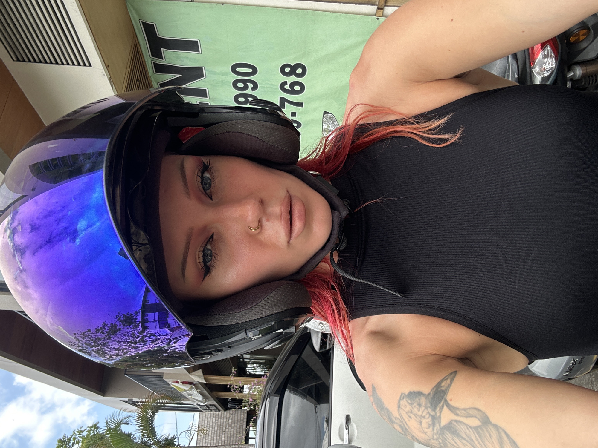A young woman with bright pink hair and a black helmet with a reflective purple visor is posing for a selfie outdoors. She is wearing a black tank top and has a nose ring and winged eyeliner. A tattoo of a bird is visible on her left arm. In the background, there is a white car, a building with a balcony, palm trees, and a green banner with black text that reads "RENT 990 768". The scene appears to be in North Kuta, Indonesia, suggesting a tropical or sub-tropical environment.