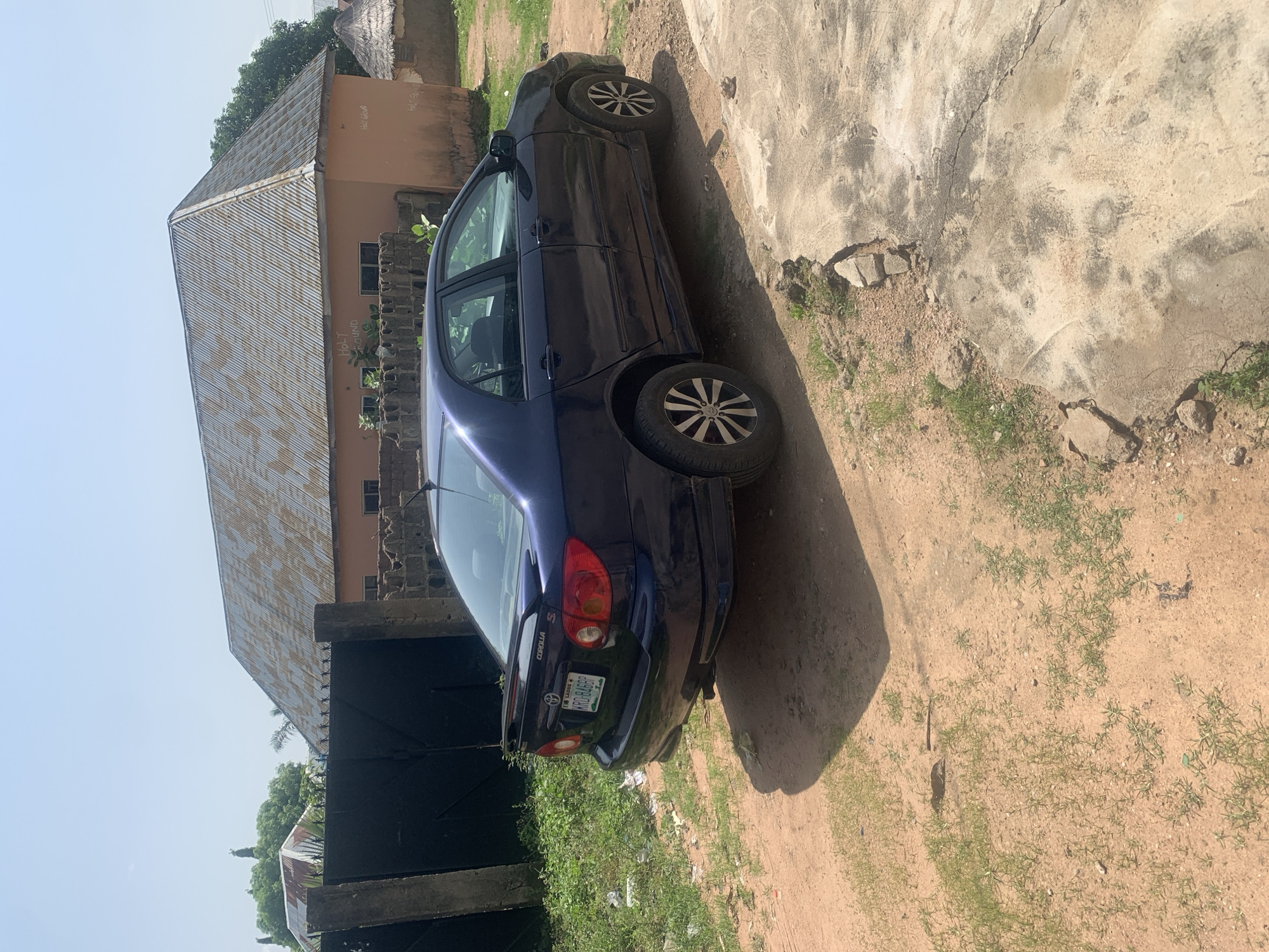 A dark blue Toyota Corolla sedan is parked on a dirt and concrete surface in front of a building with a corrugated metal roof. The car is facing away from the viewer, with its rear end visible. A license plate with the characters "KRD-846BP" is attached to the rear bumper. The building behind the car appears to be residential, with a rough, uneven roof and an orange-colored facade. A black metal gate stands to the left of the car, with a brick wall and vegetation visible beyond it. The overall environment is outdoors, under a clear, bright sky.