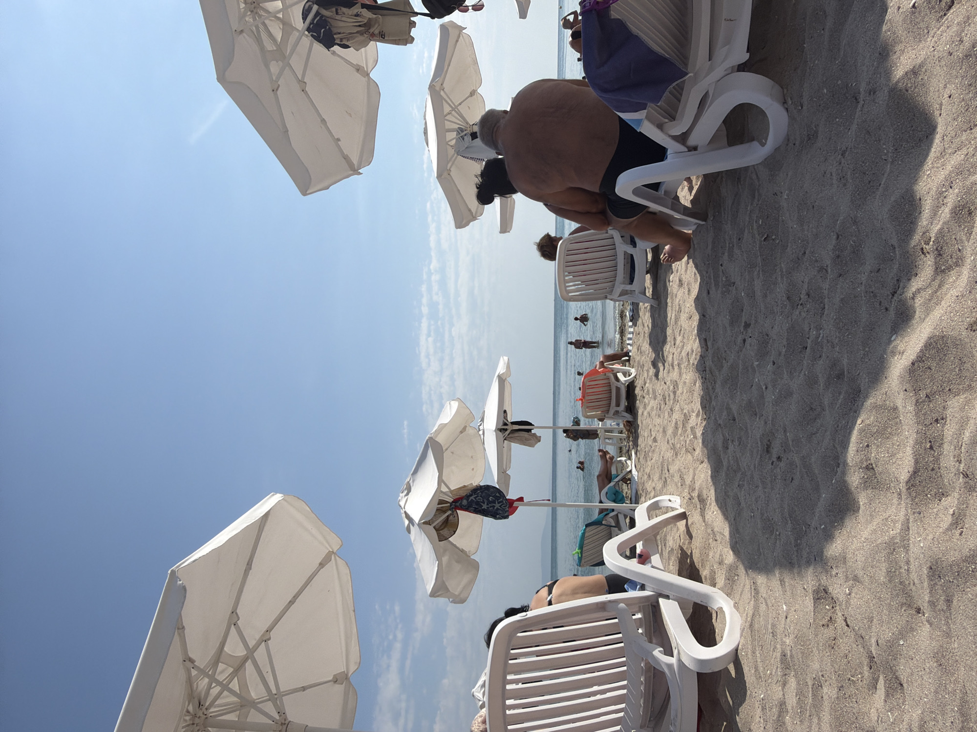The image depicts a beach scene in Pomorie, Bulgaria. In the foreground, several white plastic lounge chairs are arranged on the sand. Some are empty, while others are occupied by beachgoers. The perspective is low, looking towards the sea.
In the midground, more lounge chairs and white umbrellas are visible, providing shade. Several people are relaxing on the chairs, and some are in the water, swimming or wading. The sea is calm with a clear sky overhead.
In the background, the coastline is visible in the distance. The overall atmosphere is one of leisure and relaxation on a sunny day at the beach.