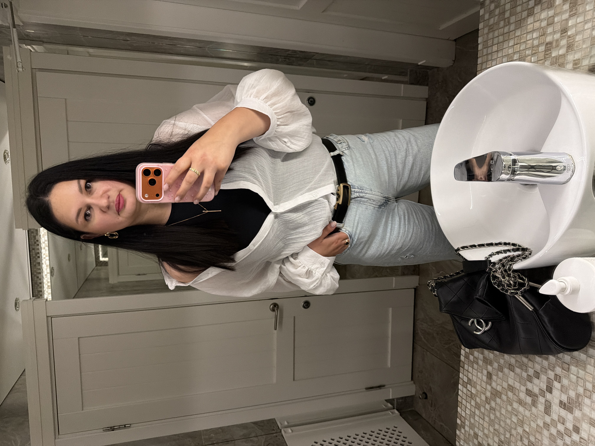 The media file shows a young woman taking a mirror selfie in a restroom. She has long, dark hair and is wearing a white, long-sleeved blouse, a black camisole, light-wash jeans, and a black belt with a gold buckle. She is also wearing gold hoop earrings and a ring on her right hand. She is holding a pink iPhone in her right hand, which is visible in the reflection. A black quilted handbag with a silver chain strap is placed on top of the sink basin. The restroom has tiled walls and doors, and a white sink with a chrome faucet. The overall mood appears casual and confident. There is no visible text in the media file.