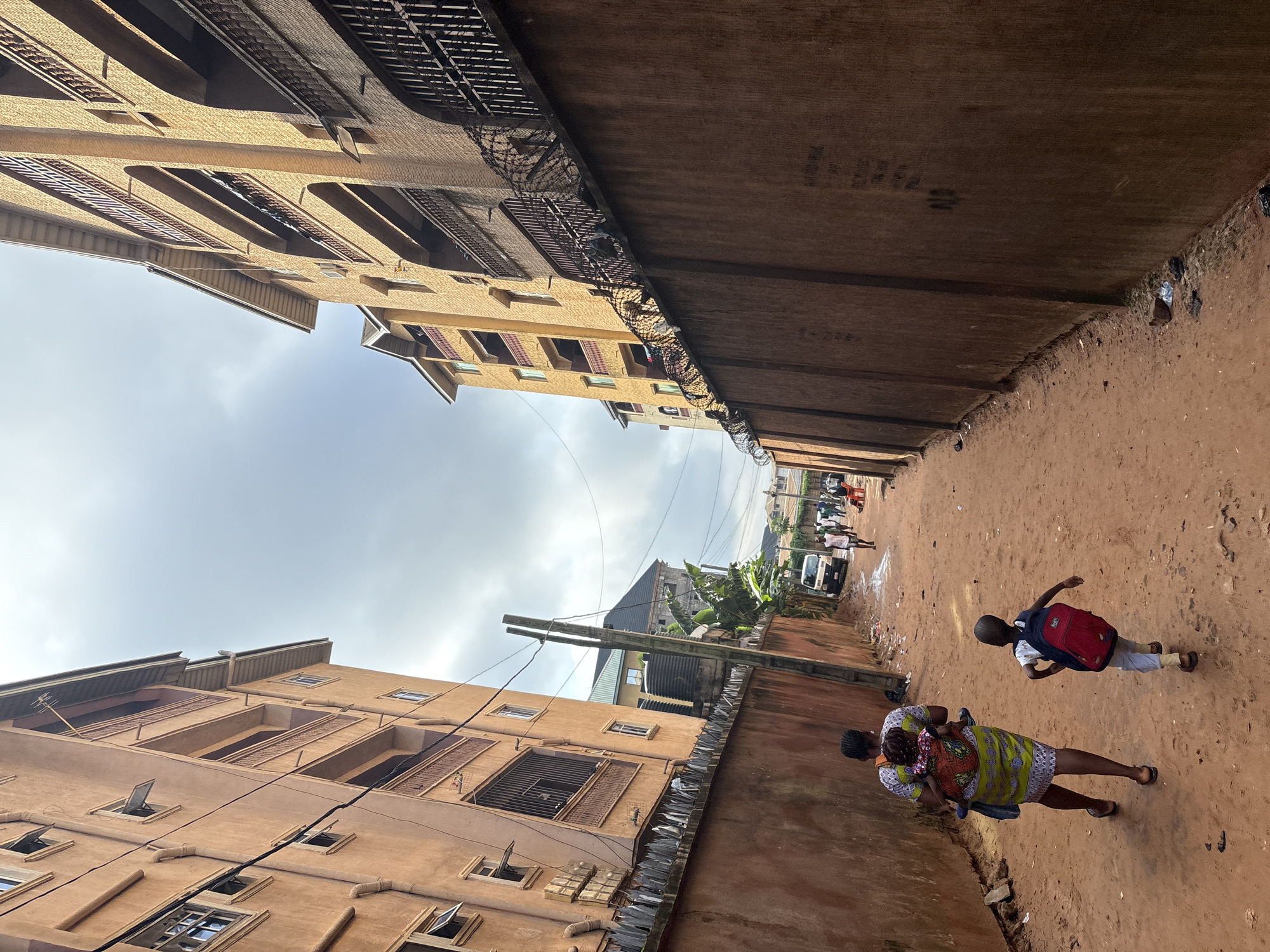 A low-angle, full shot shows a dirt pathway between two large, multi-story residential buildings in Onitsha, Nigeria. The building on the left is a light orange color, with balconies on each of its upper floors. The building on the right is a yellowish-brown color, also featuring balconies and arched windows. The pathway is composed of reddish-brown dirt and is lined by a tall, reddish-brown wall on either side. Barbed wire is visible atop the wall on the right.
In the foreground, a woman walks away from the camera, carrying a baby on her back, who is wrapped in a colorful fabric. Next to her, a young boy, also walking away, wears a red backpack. In the background, a white van is parked on the pathway, and several other people, some appearing to be walking and some sitting, are visible further down the path. The sky above is overcast with a light gray color.
