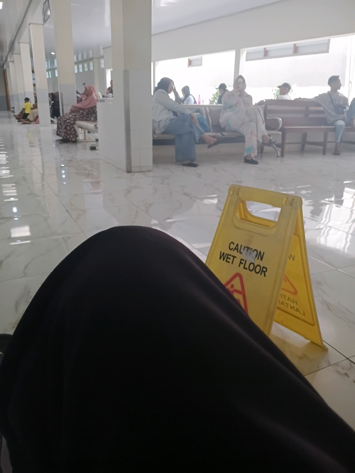 The image depicts an indoor waiting area, likely a public building such as a clinic or hospital, with a polished marble floor. Several people are seated on benches in the background, some appearing to be patients or visitors. A yellow "Caution Wet Floor" sign is prominently displayed in the foreground, indicating a potential hazard. The overall lighting suggests an indoor environment with overhead illumination. The specific location of Purwodadi, Indonesia, cannot be determined from the visual information present in the image.