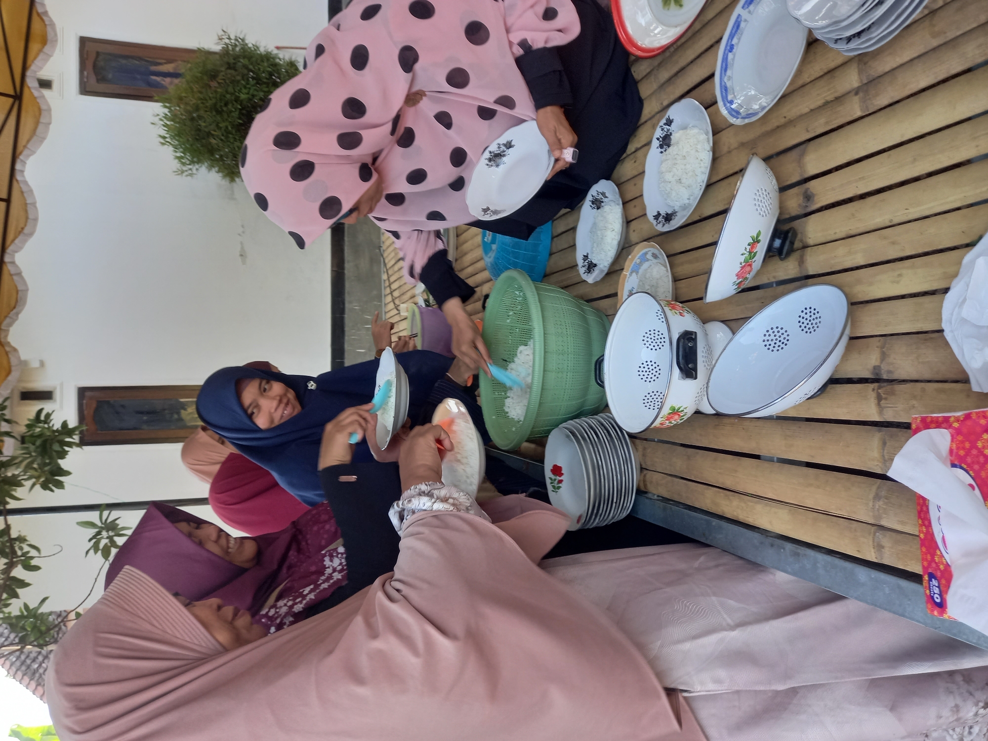 In Tempos, Indonesia, a group of women are gathered outdoors around a bamboo table, serving rice. Four women are clearly visible, all wearing headscarves. One woman in a pink headscarf with black polka dots is using a spoon to serve rice onto a plate. To her left, a woman in a blue headscarf is also serving rice. Another woman, wearing a maroon headscarf, is smiling and looking at the woman in blue. A fourth woman, in a light pink headscarf, is holding a plate with rice. A large green colander filled with rice is on the table, along with several plates of rice, stacked plates, and a box of tissues. The background shows a white wall and some greenery, suggesting an outdoor or semi-outdoor setting.