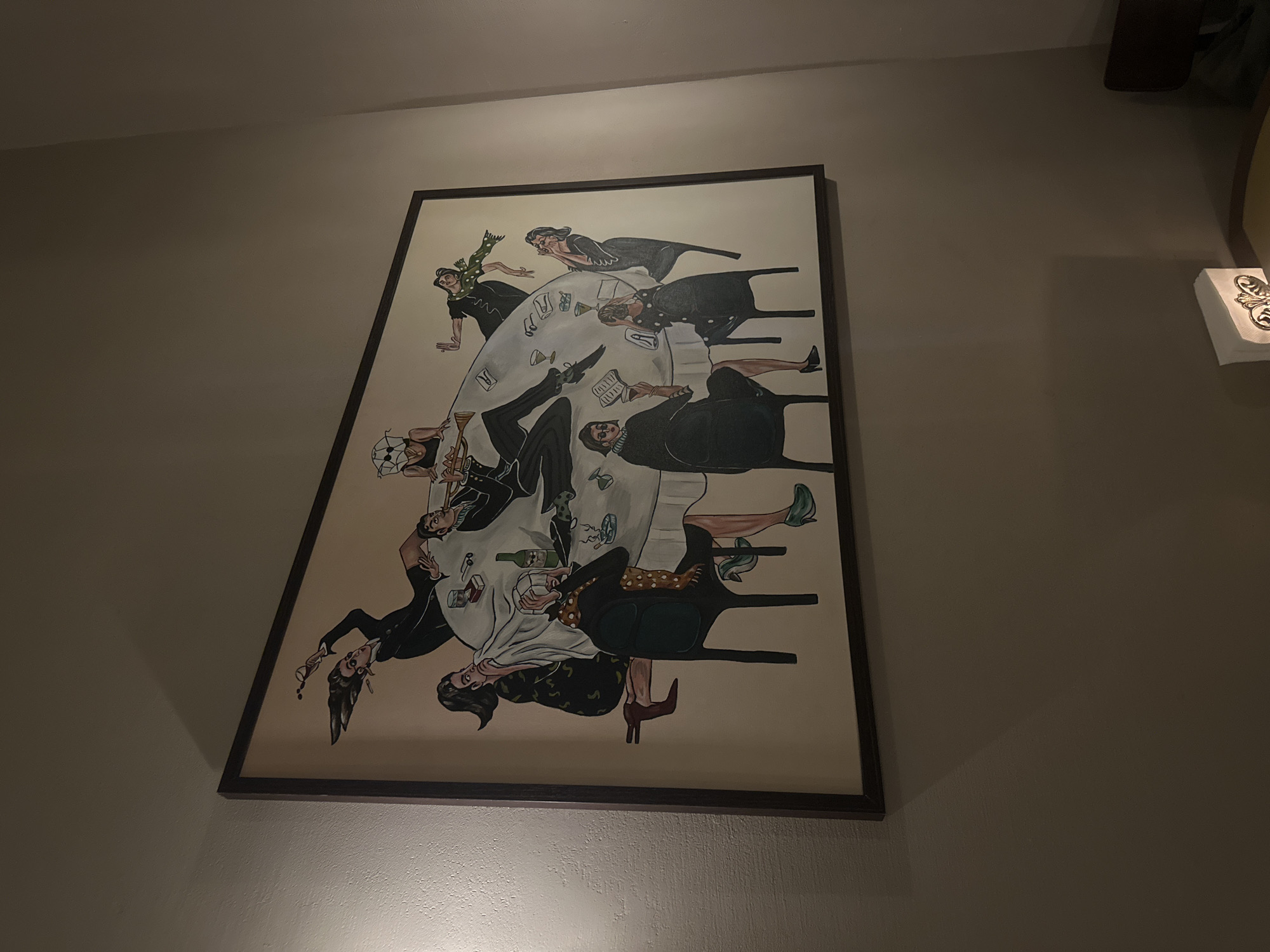 A medium shot depicts a framed painting hanging on a light tan or gray wall. The artwork appears to be a gathering of approximately nine stylized figures seated or leaning around an elongated oval table covered in a white tablecloth. The figures are dressed in black dresses or suits with various accessories. The table is scattered with objects like glasses, bottles, papers, and books. The style of the figures and objects suggests an art deco or surrealist influence. To the right of the framed painting, a partial view of a shelf with a decorative object resting on it can be seen. The wall appears to be smooth and evenly lit. This painting is located in Yerevan, Armenia.