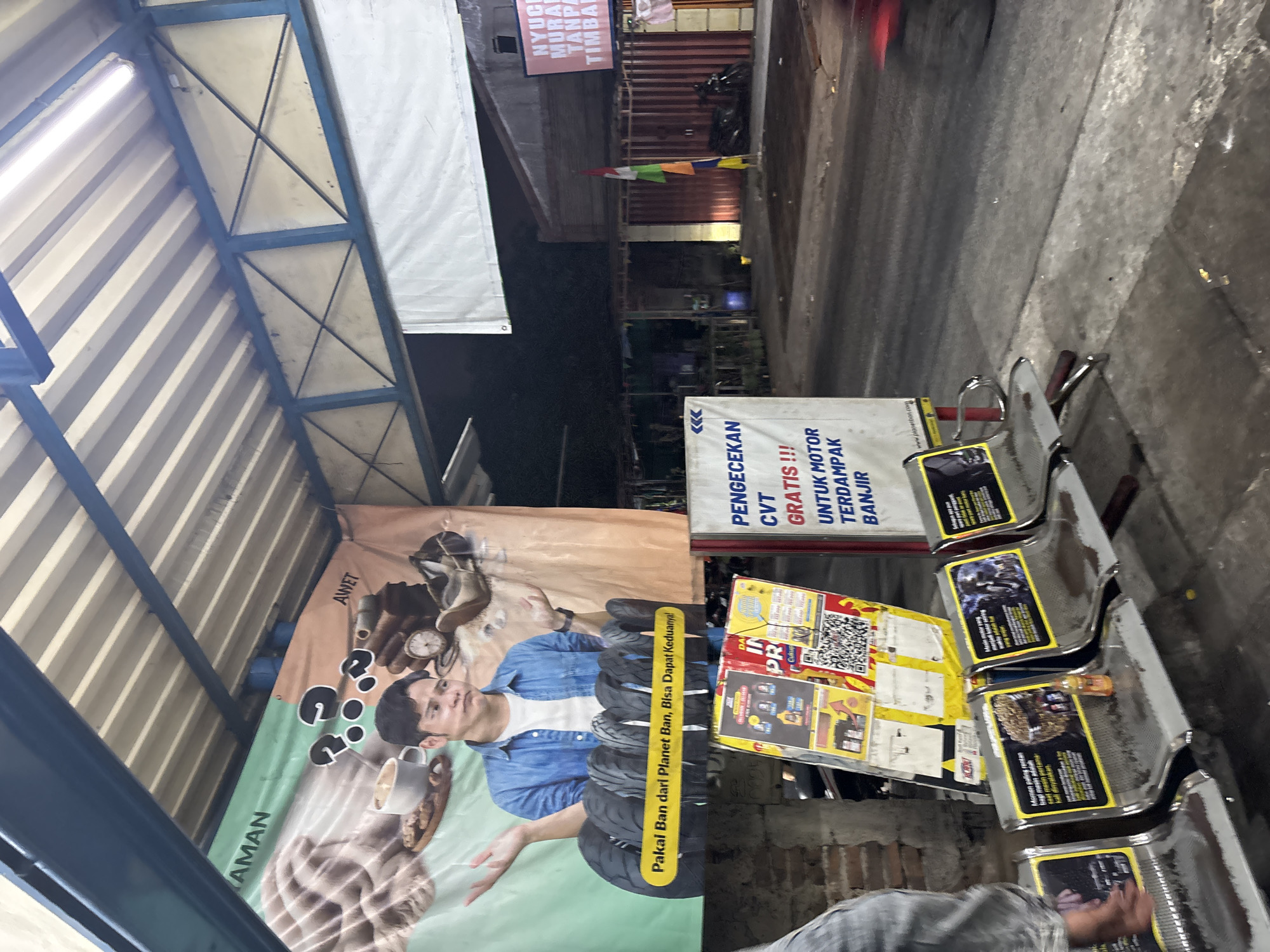 The image depicts an outdoor waiting area, likely a bus stop or transit hub, in North Jakarta, Indonesia. A prominent banner features a man with a perplexed expression, gesturing towards a display of tires and text in Indonesian that translates to "Wear Tires from Planet Ban, You Can Get Both!" Below this banner, a row of empty metallic waiting chairs is visible, some adorned with advertisements. One advertisement on a chair shows a close-up of a helmeted motorcyclist. Another displays an image of gravel and the text "The most enjoyable moment for riding a motorcycle is when you first experience it. You must try it." To the right, a vertical sign advertises "CVT CHECK FREE!!! FOR MOTORCYCLES WITHOUT CLUTCHES." The background shows a street scene at dusk or night, with buildings and a parked motorcycle. The overall ambiance suggests a commercial or service area, possibly near a motorcycle repair shop or a place offering related services.