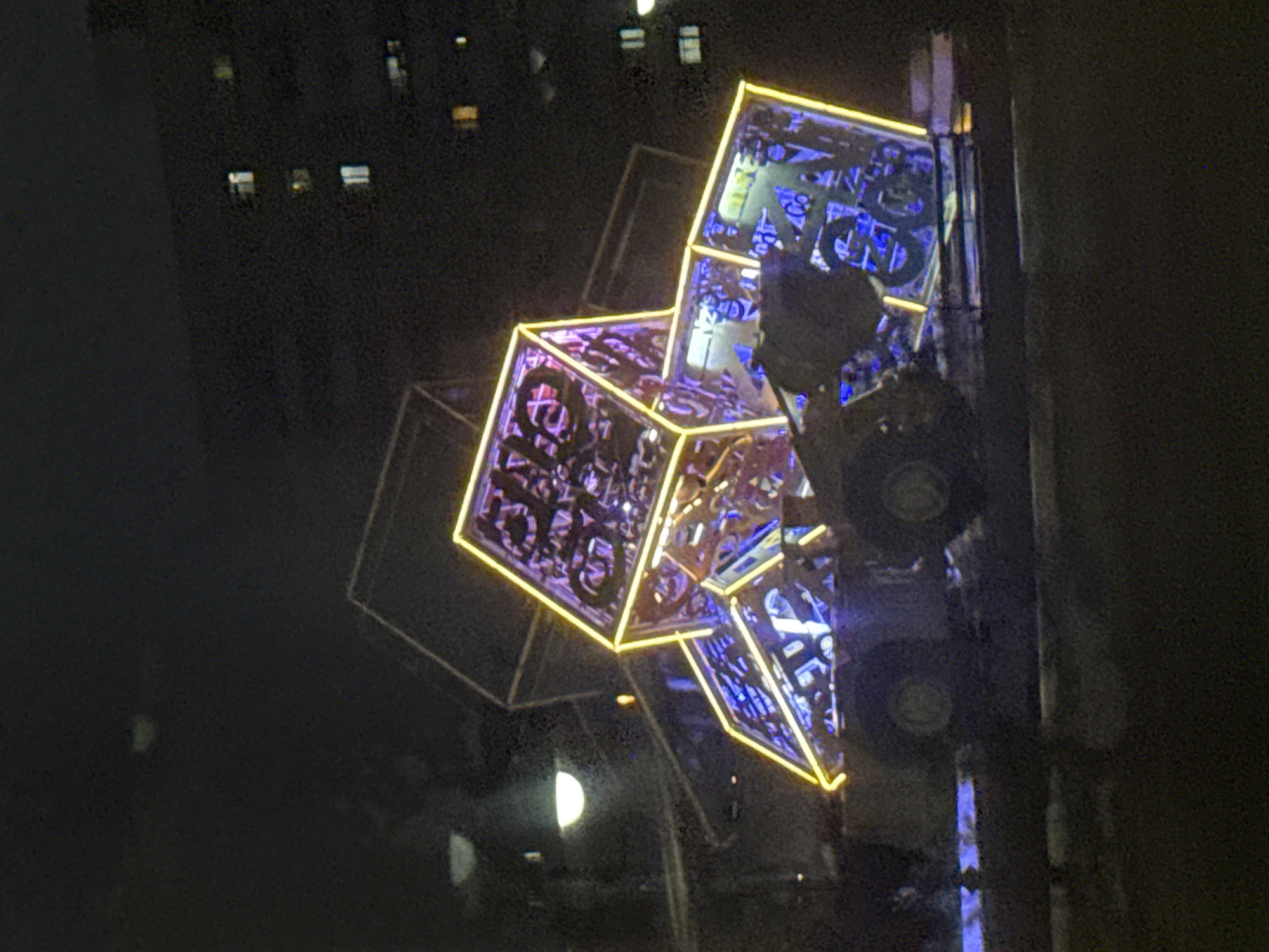 A nighttime exterior scene in Norilsk, Russia, features a large, abstract sculpture composed of illuminated, geometric shapes. The sculpture, possibly an art installation, is lit with bright yellow and blue lights, creating a vibrant contrast against the dark sky. The shapes are angular and appear to be made of metal or glass, with intricate patterns visible within them. The sculpture is supported by what appears to be heavy machinery, possibly a loader or a similar industrial vehicle, with large wheels visible at the base. In the background, the facade of a multi-story building is visible, with several windows illuminated, suggesting occupancy. The ground appears to be paved, with some light reflecting from the sculpture onto the surface. The overall impression is one of a modern, urban setting at night, possibly during a festival or exhibition, given the prominent art installation.