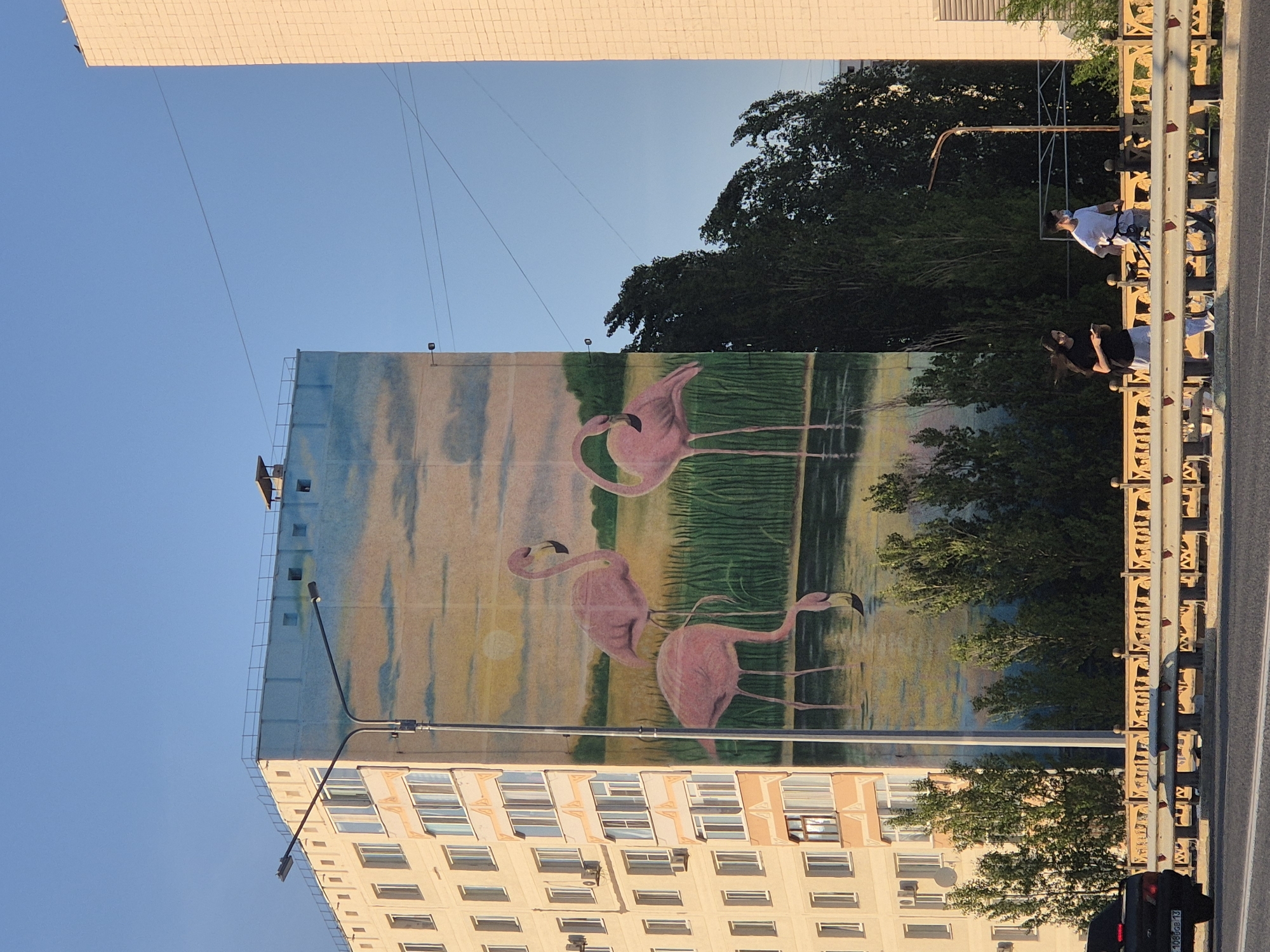 A building in Kokshetau, Kazakhstan, displays a mural of three pink flamingos in a waterscape. The lower portion of the building is tan with numerous windows. A street light is positioned near the building. A pedestrian overpass runs parallel to the building. Two people stand on the overpass. A black car is on the street to the right. The sky is blue.