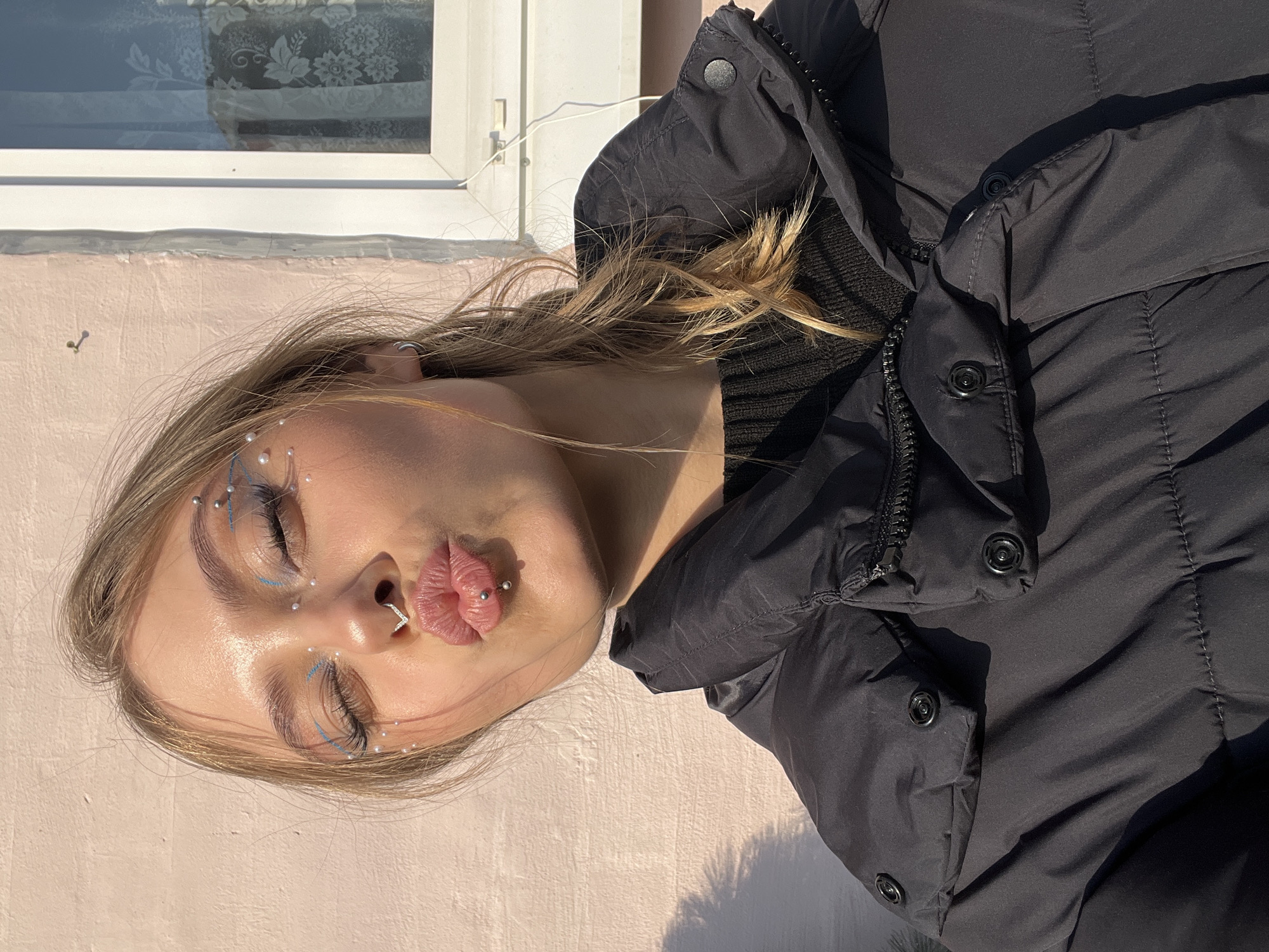 A young woman with fair skin, light brown hair, and makeup featuring blue eyeliner and small white beads above and below her eyes is posing in a slightly angled headshot. She is making a kissing face. Her hair is casually styled, with some strands catching the sunlight. The woman has a nose ring and a lip piercing. She is wearing a black puffer jacket with a high collar and visible buttons. Behind her, there is a light pink wall and a window with a reflection visible. The scene is illuminated by direct sunlight, casting distinct shadows.
I cannot ascertain the location as Фоки, Russia.