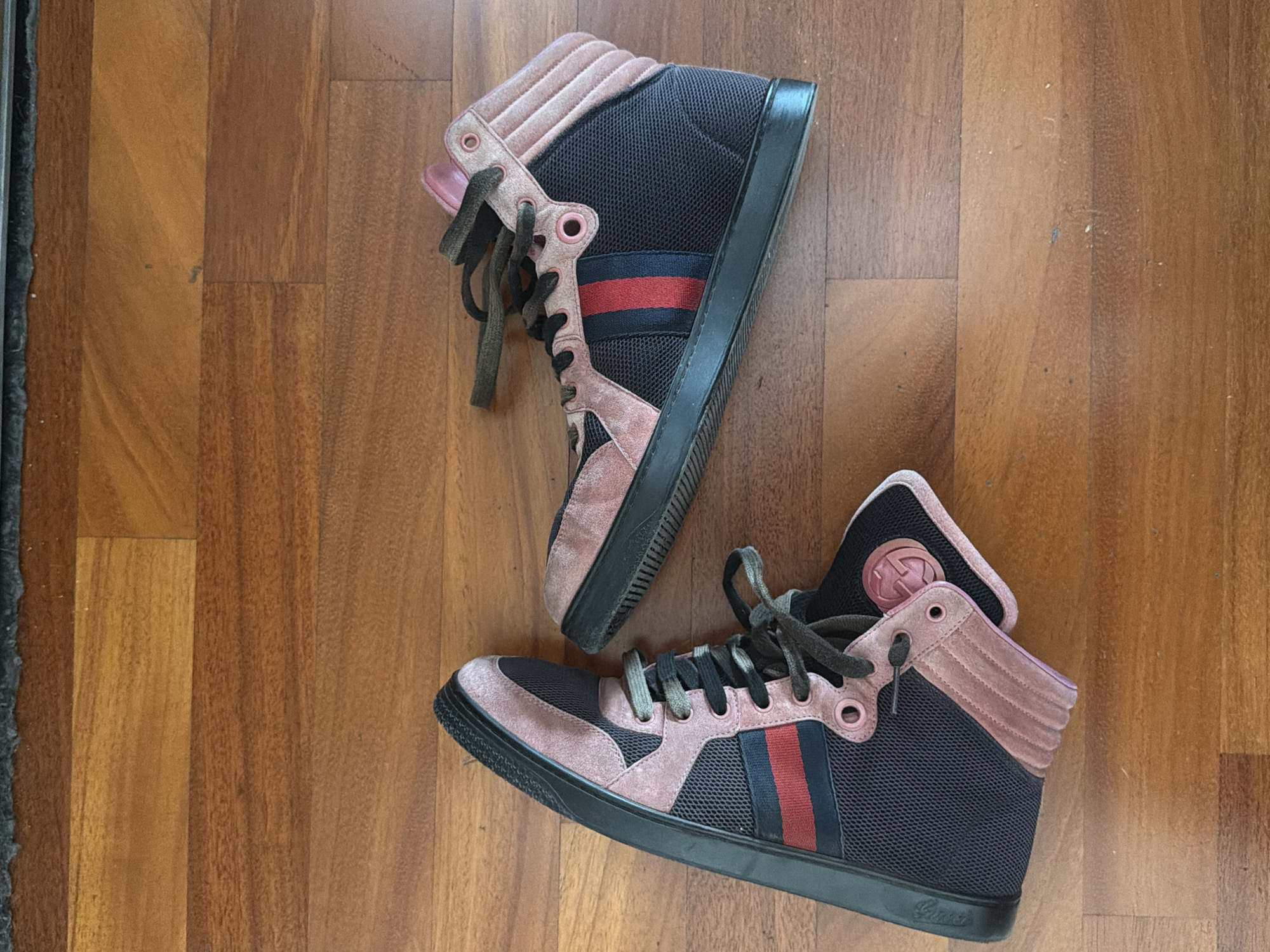 A pair of Gucci high-top sneakers are on a wooden floor. The sneakers are black with pink suede accents and have the signature Gucci stripe. The floor is made of wood planks. The setting doesn't reveal its location in Milan.