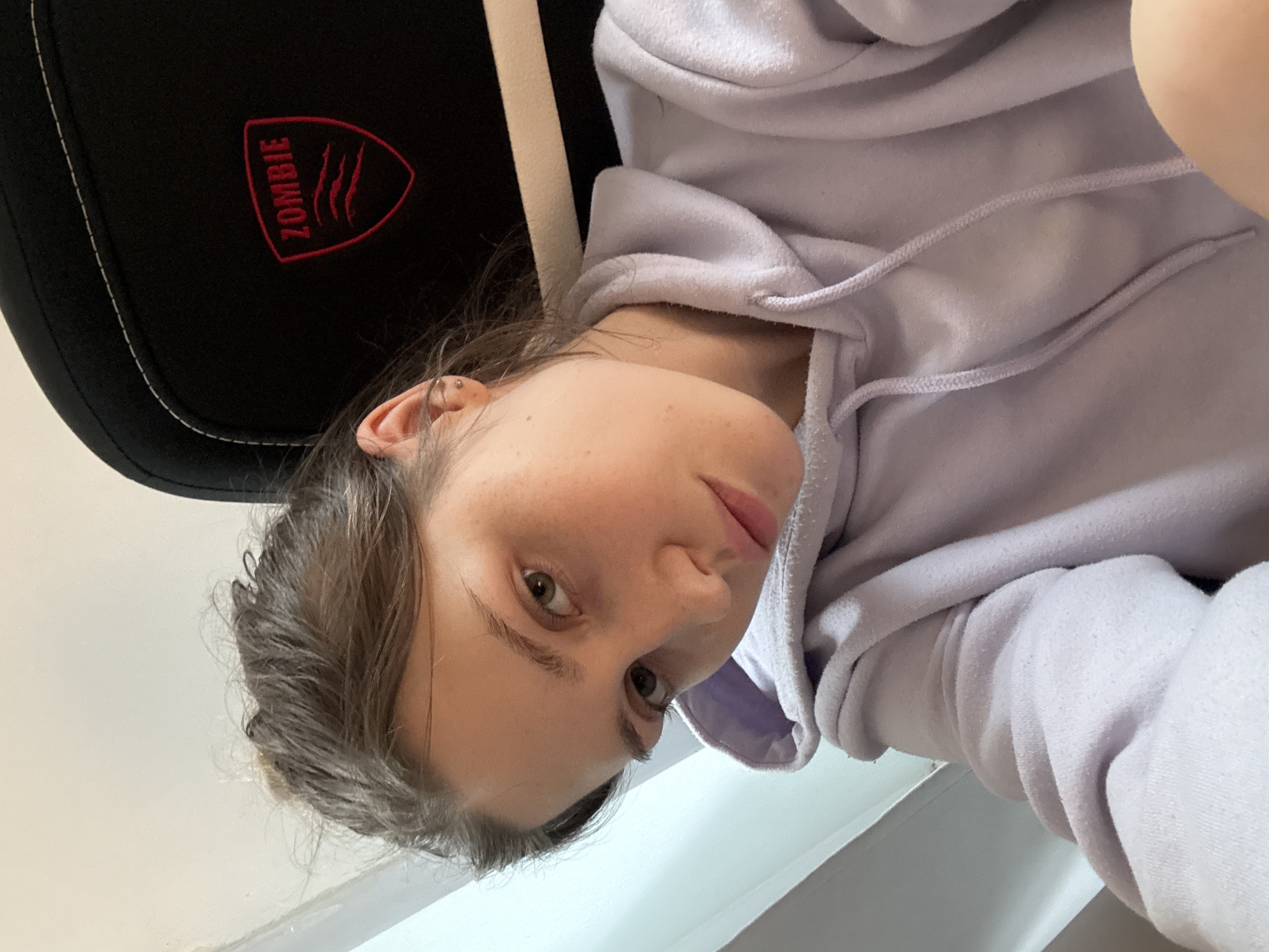 A close-up portrait shows a young woman looking directly at the camera, with her head tilted significantly to her right. She has light brown hair, and striking light-colored eyes, possibly hazel or green. A small silver helix piercing is visible on her left ear. She is wearing a soft, pastel purple or lavender hoodie, with its drawstrings clearly visible around the neckline.
The woman appears to be seated in a black gaming chair, which is partially visible behind her head and shoulder. The chair features a prominent red embroidered logo on its headrest. The logo consists of the word "ZOMBIE" in capital letters, accompanied by a stylized three-claw mark design enclosed within a shield shape. White stitching outlines parts of the chair. The background is mostly a plain, light-colored surface, suggesting an indoor setting with natural light. The overall mood is relaxed and casual. The image captures a moment, likely a selfie or a close-up snapshot, set in Saint Petersburg, Russia.