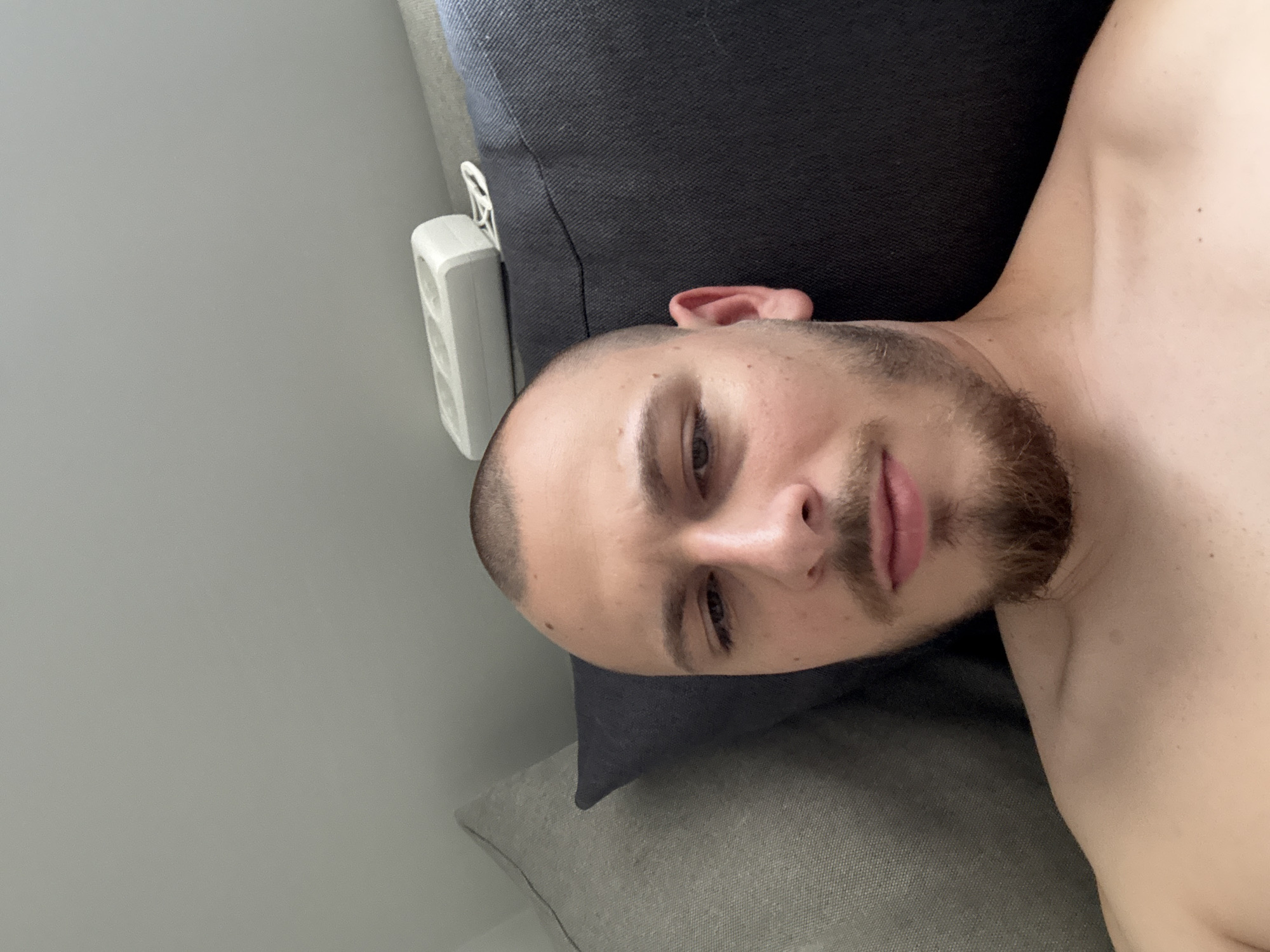 A close-up, slightly angled, full-face image features a man with short, buzzed brown hair and a brown beard. He is lying down, possibly on a sofa, with his head resting on a light grey pillow. The sofa behind him is dark grey. A white electrical outlet is visible on the wall to the left, next to the sofa. The man's gaze is directed toward the camera. He appears to be shirtless. There is no discernible information to suggest the image was taken in Stara Zagora, Bulgaria.