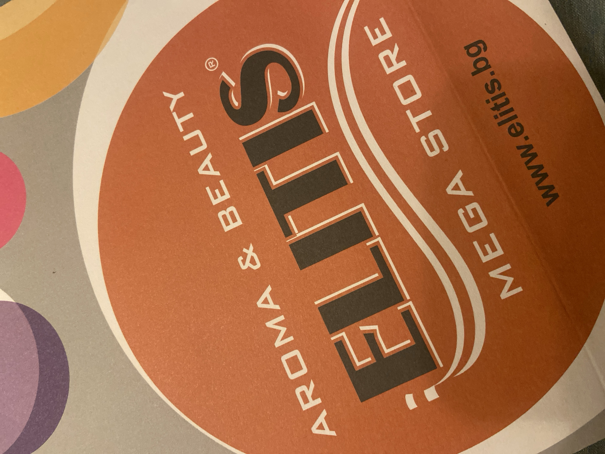 The image is a close-up of a circular sticker or label, likely for promotional purposes. The background of the sticker is a vibrant orange. At the top, in white uppercase letters, is the text "AROMA & BEAUTY". Below this, prominently displayed in large, dark gray and white font, is the word "ELITIS". A small registered trademark symbol (®) is visible to the right of "ELITIS". Beneath the large text, in white uppercase letters, is the phrase "MEGA STORE". The bottom of the sticker features the website address "www.elitis.bg" in black text. The sticker has a curved white element running under the "ELITIS" text, resembling a stylized wave or ribbon. The overall impression is of a branding or advertising element for a store. The image does not contain any information about a specific city or location.