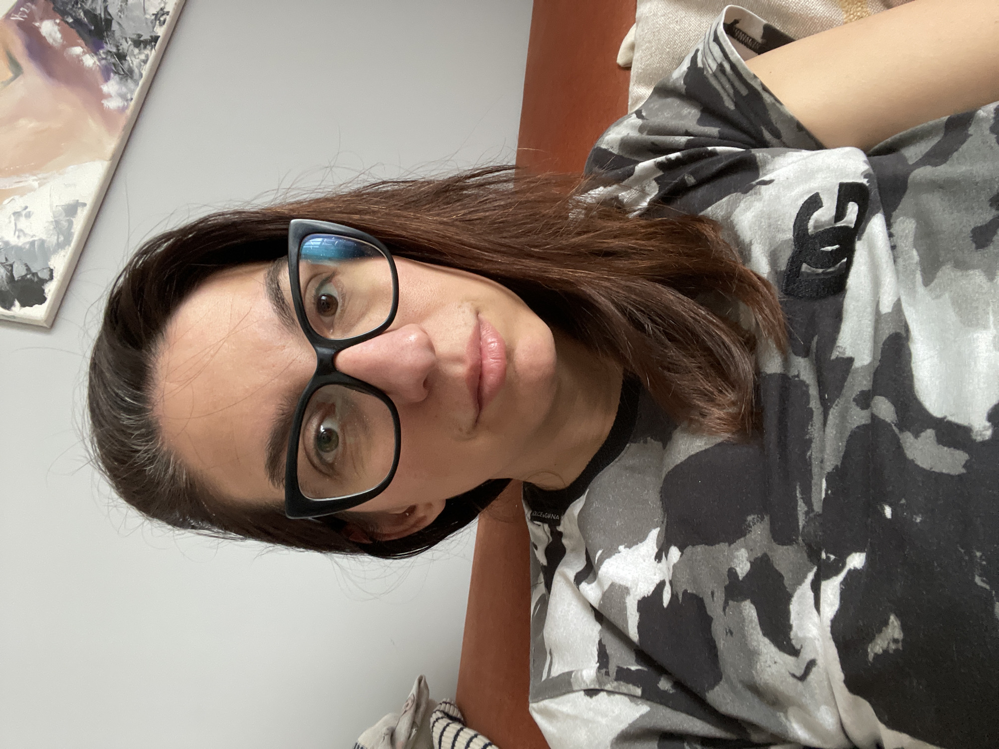 A woman with brown hair and glasses is indoors, lying on a brown or orange surface. She is wearing a grey and black camouflage-patterned shirt. A painting is visible in the background on a white wall. No location information.