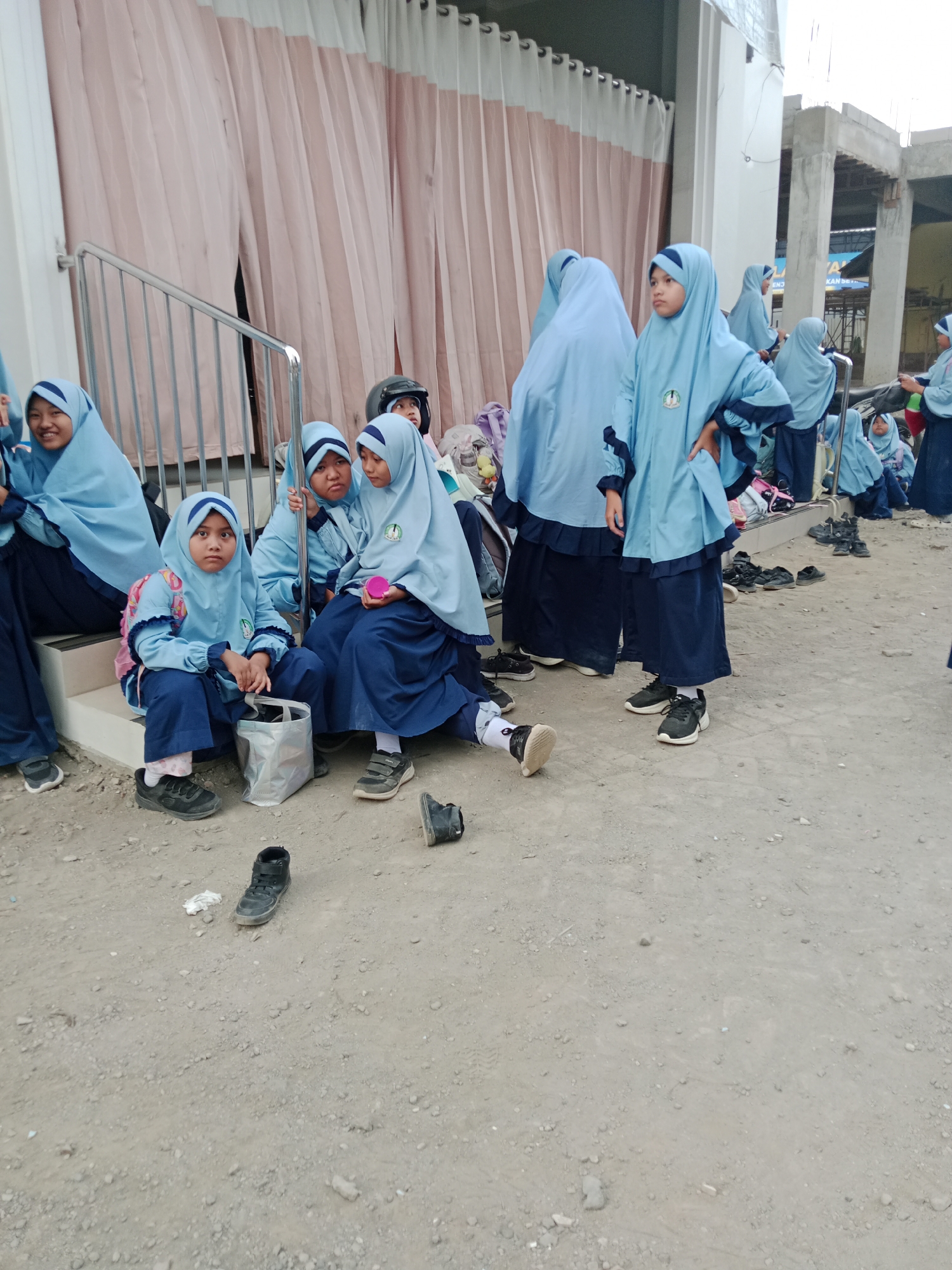 The image depicts a group of young girls wearing light blue hijabs and dark blue uniforms, suggesting they are students. They are seated on concrete steps and the ground in an outdoor area. The environment appears to be under construction, with exposed concrete structures and scaffolding visible in the background, along with draped fabric that could be temporary covering. A metal railing runs along the steps. Several pairs of black shoes are scattered on the ground, indicating that the girls may have removed them. One girl in the foreground is looking directly at the camera. The scene is likely located in Kediri City, Indonesia, given the context.