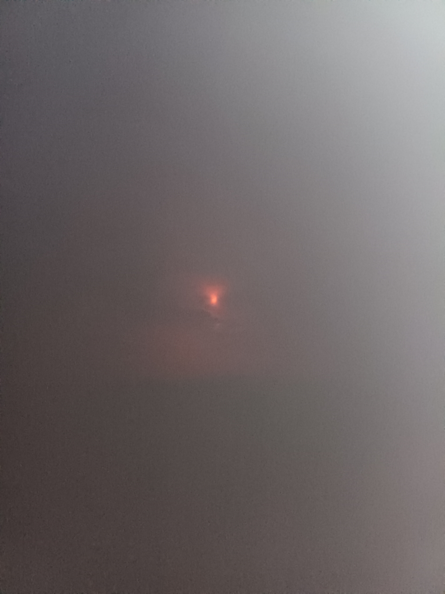 The image shows a hazy sky with an intense, reddish-orange circular light in the center, likely the sun obscured by heavy fog or smoke. The surrounding sky is a uniform gray, indicating low visibility. There are no discernible objects or people in the scene. The image appears to be taken outdoors in an urban or suburban environment, though the haze obscures any specific landmarks.